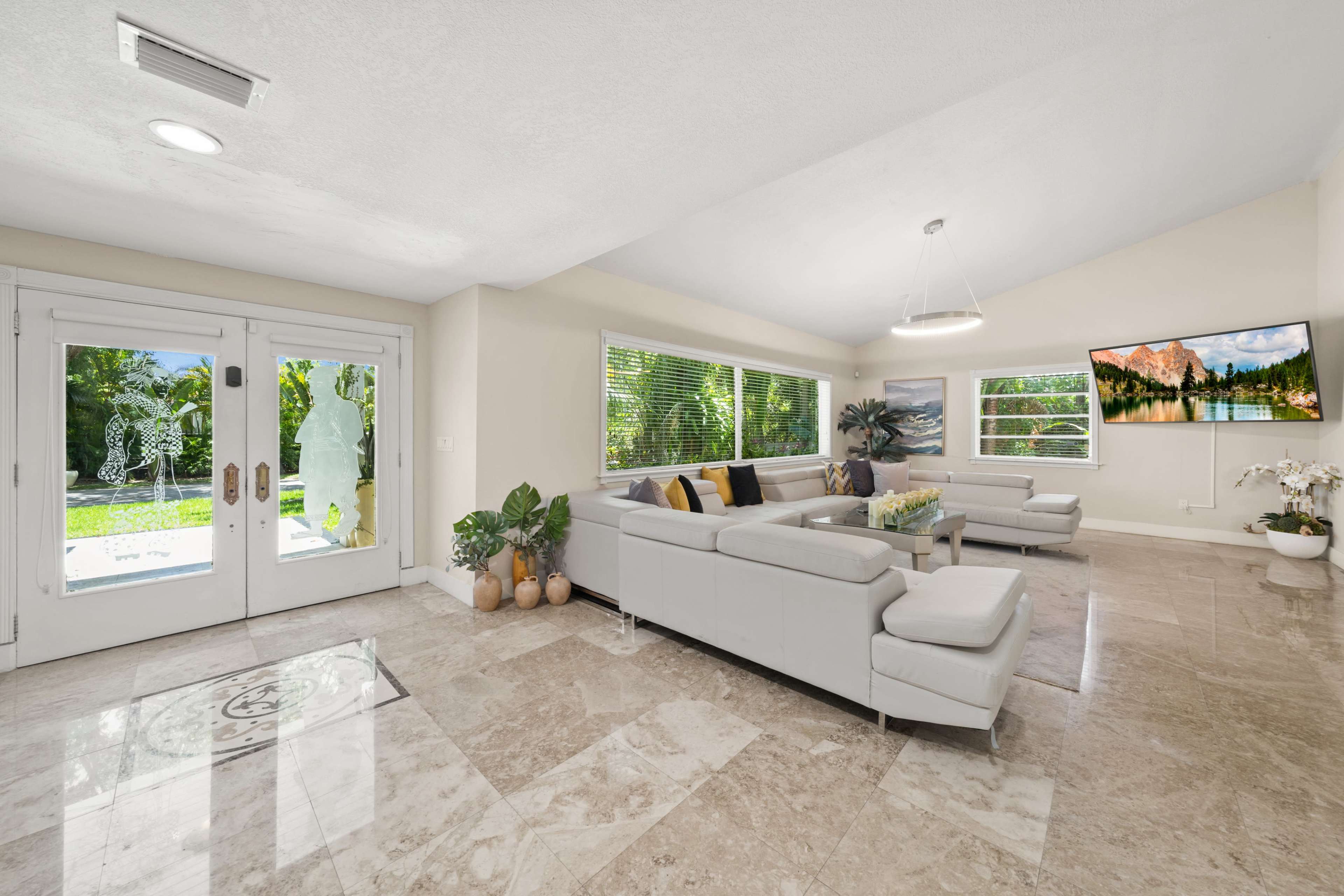 A bright living room features a sectional sofa, a wall-mounted TV, and large windows overlooking a garden.