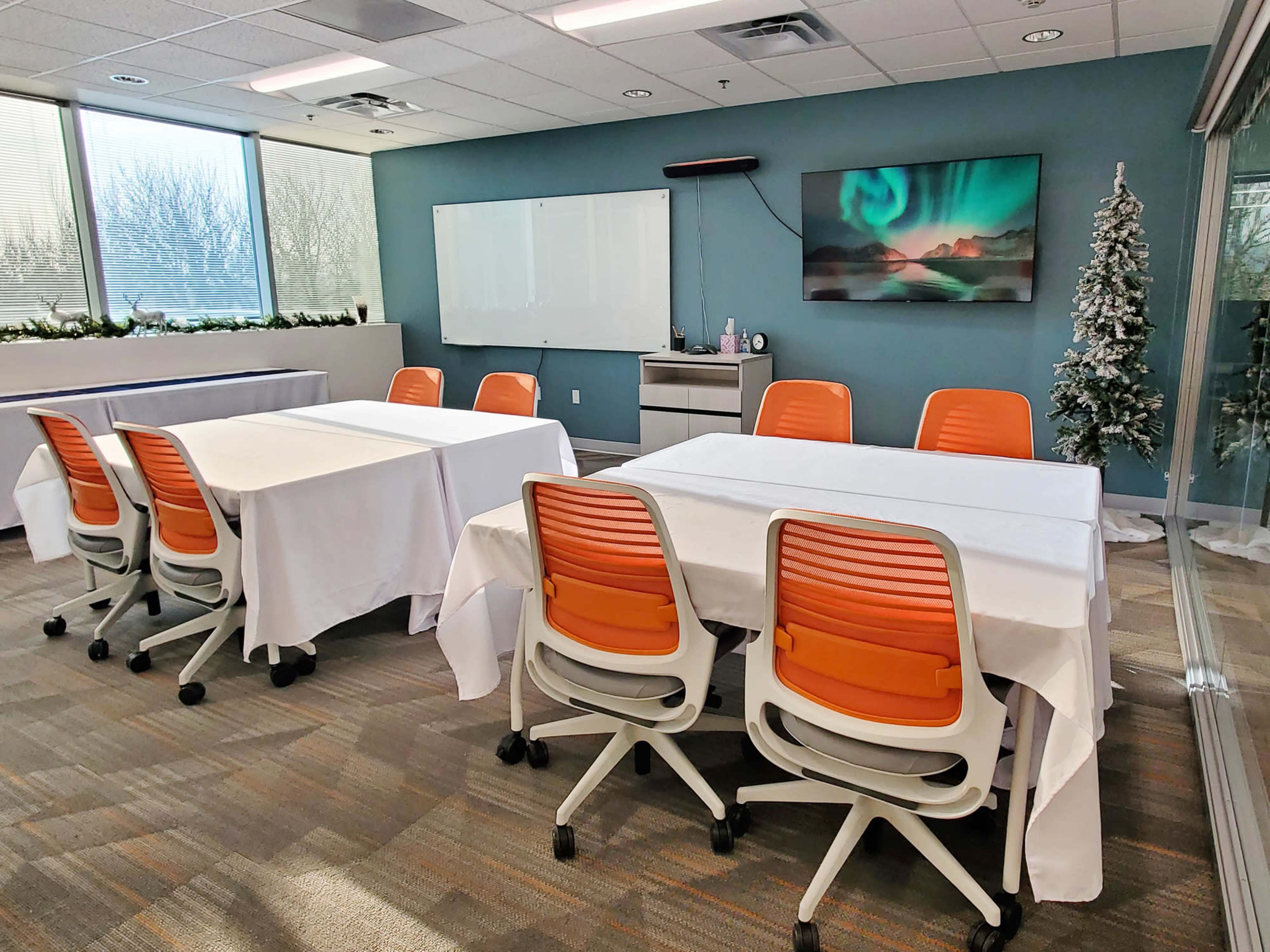 A conference room features several tables covered with white tablecloths and orange chairs, along with a large screen displaying a landscape image and a decorative tree in the corner.
