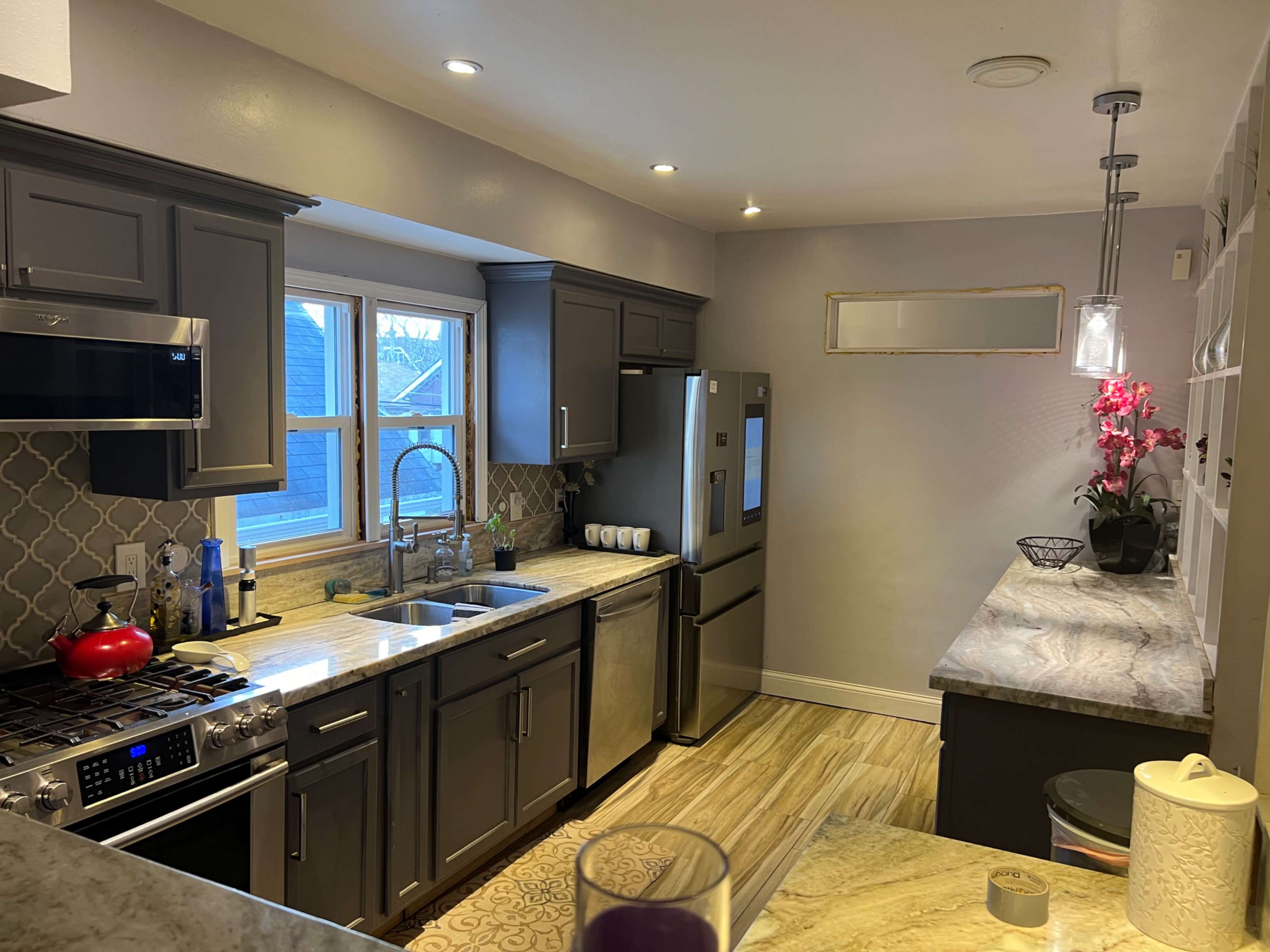 The image shows a modern kitchen with gray cabinets, stainless steel appliances, and a marble countertop, illuminated by natural light from two windows.