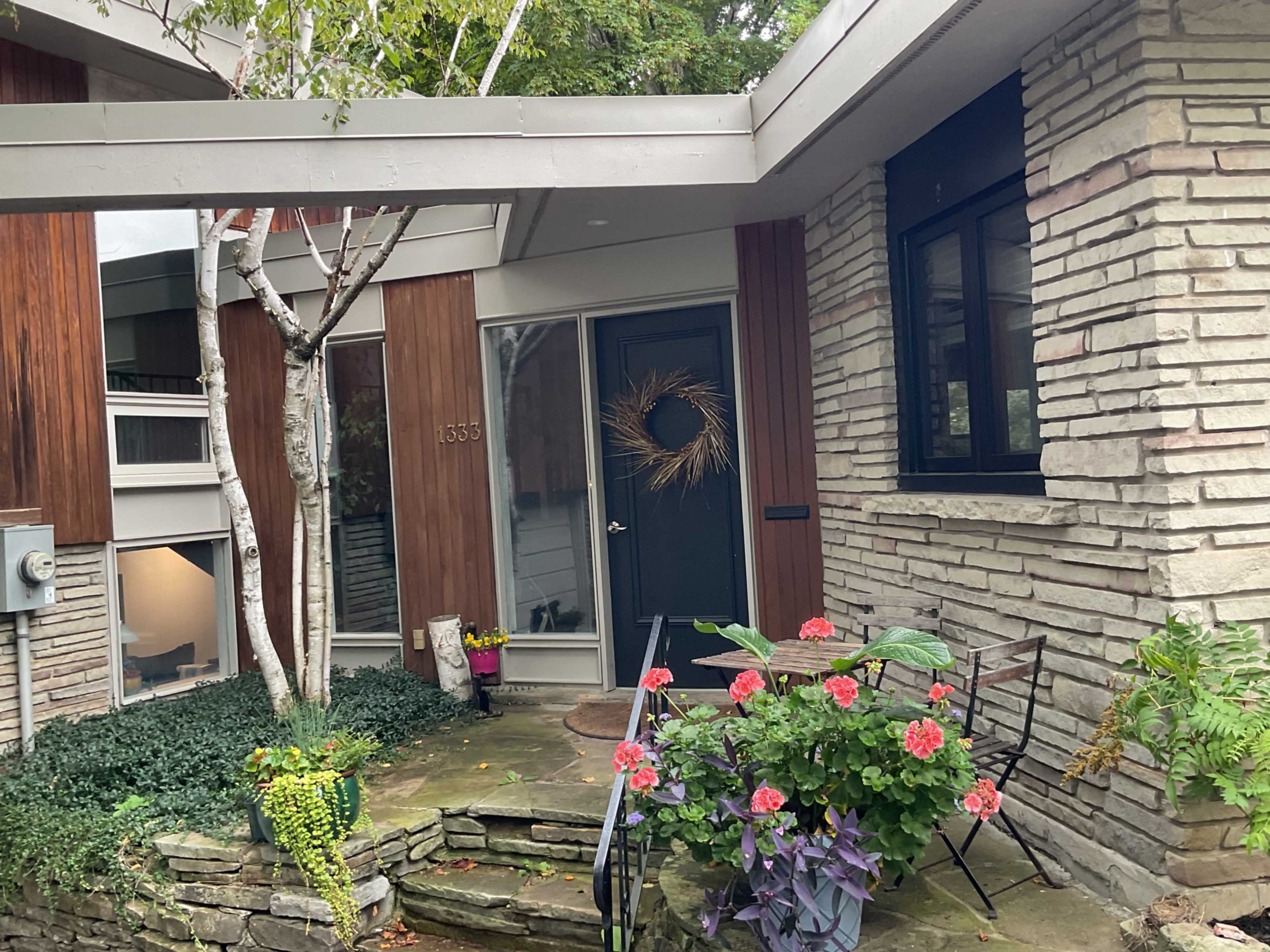 The image shows a modern house entrance with wooden paneling, a stone facade, a front door decorated with a wreath, and vibrant flower planters beside a small stone pathway.