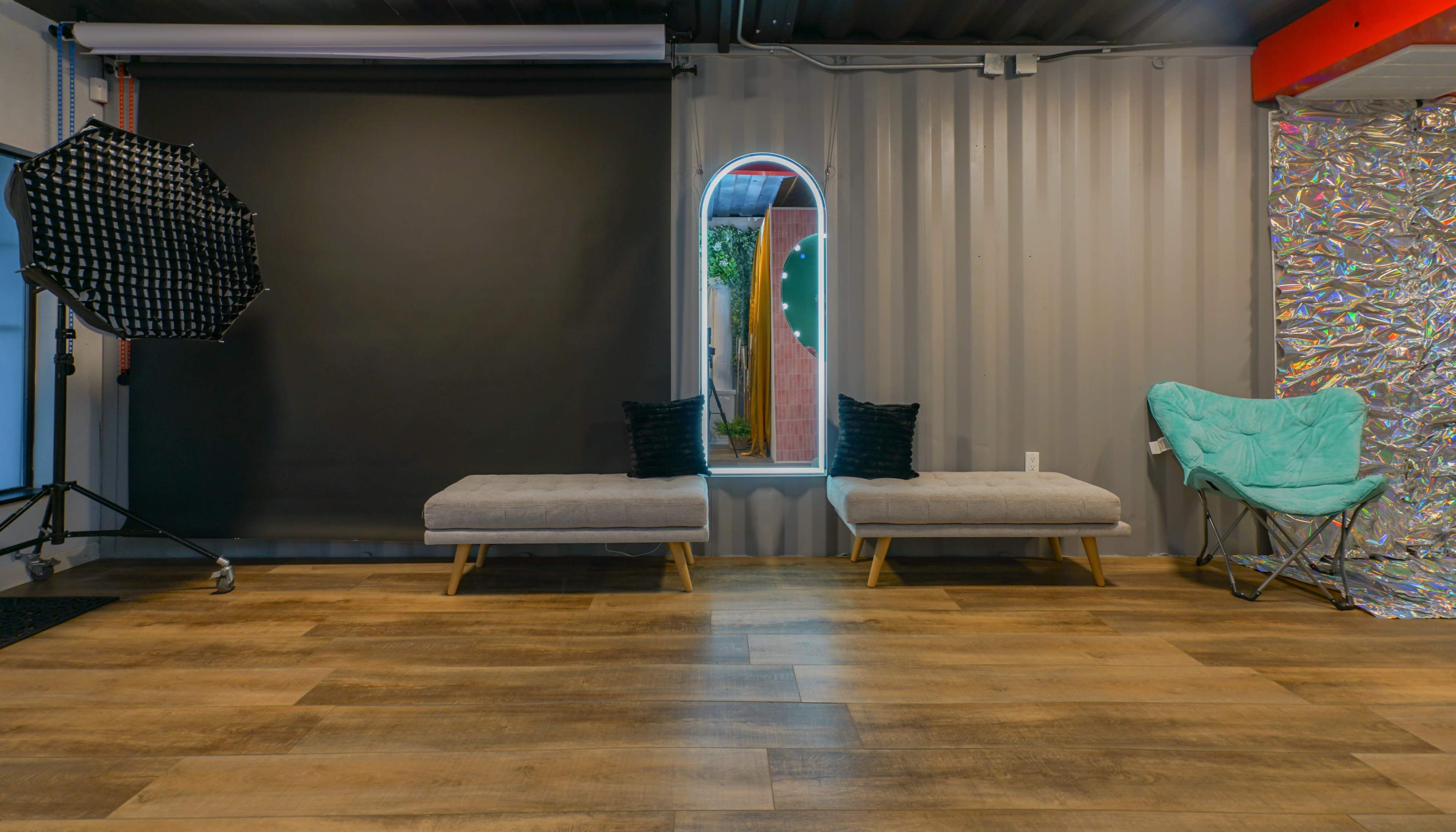The image shows a contemporary photography studio with a gray backdrop, two gray benches, and a turquoise chair against a wall of corrugated metal.