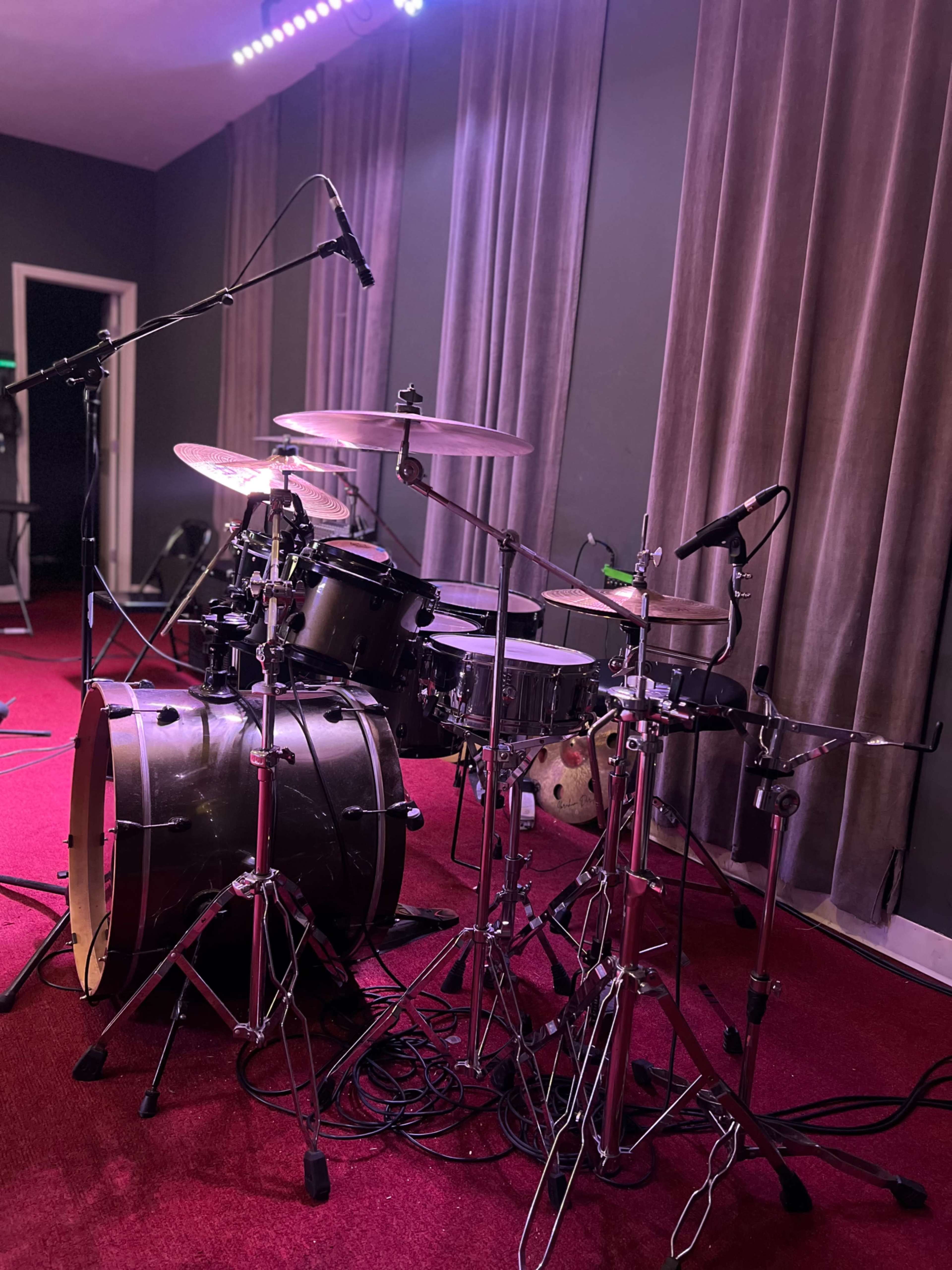 The image shows a drum set arranged with various components, including a bass drum, snare, cymbals, and microphones, set up on a red carpeted stage with curtains in the background.