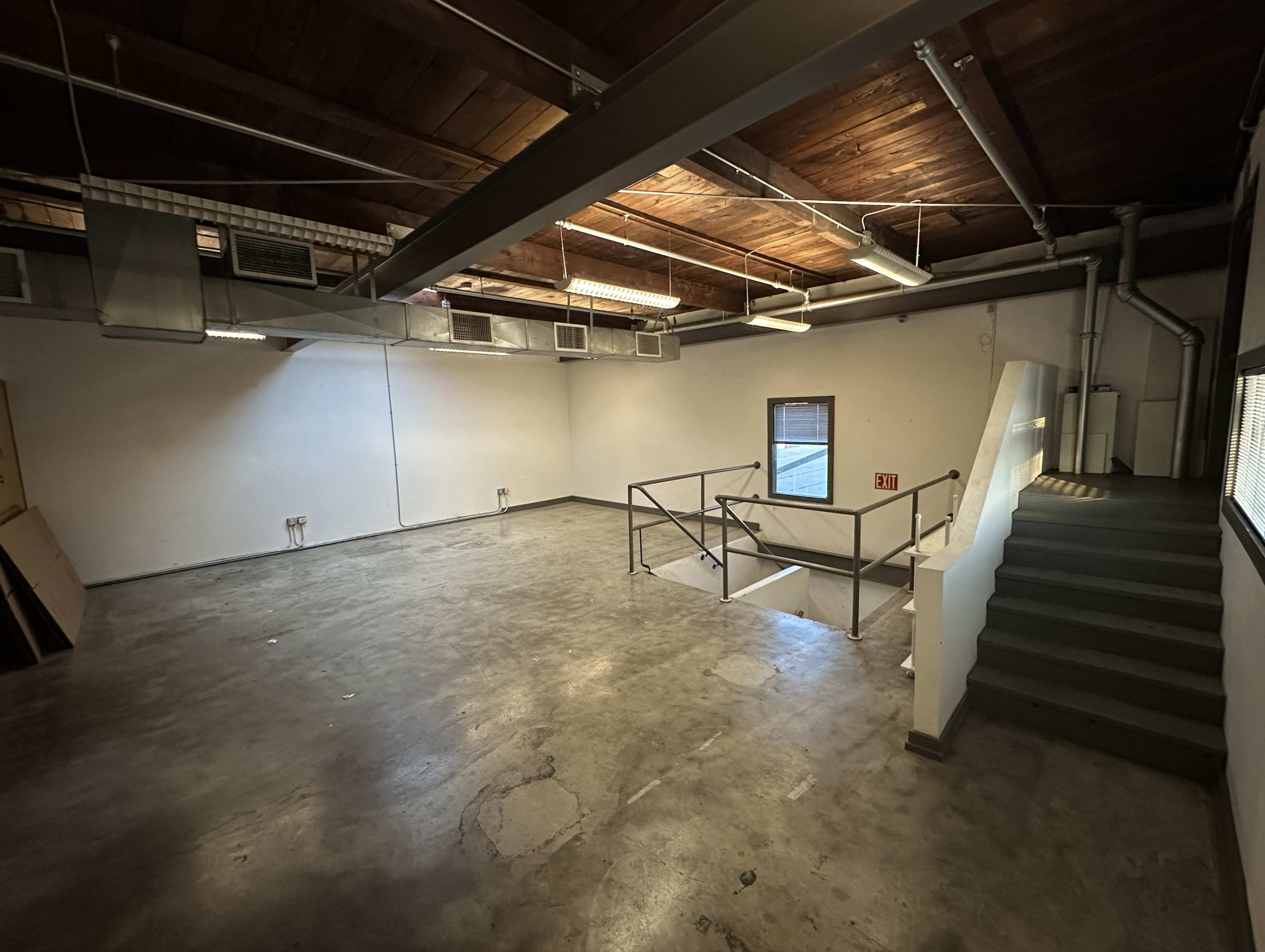 The image shows an empty industrial-style room with a concrete floor, wooden ceiling, and a staircase leading to a small upper level.