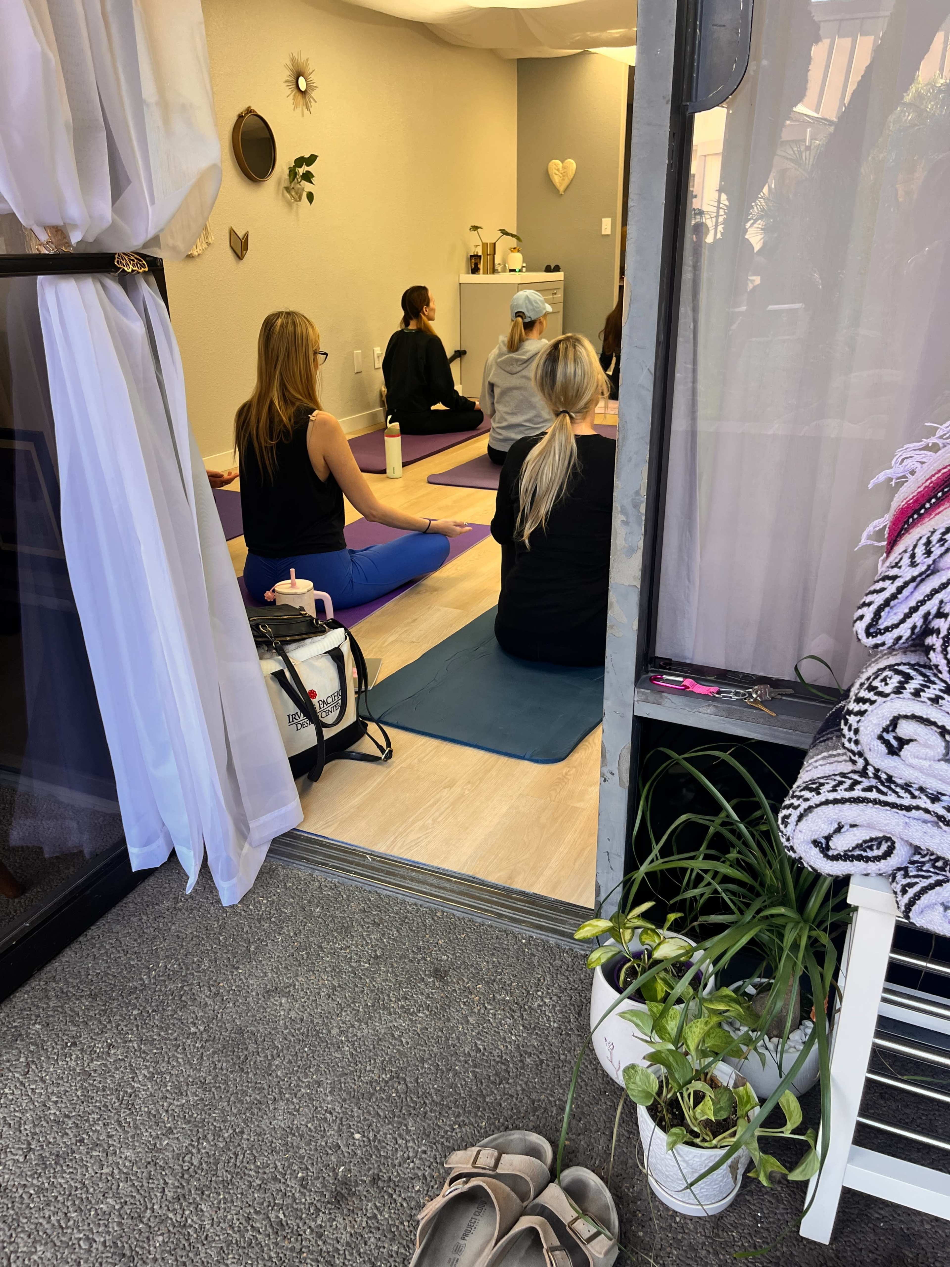 A group of people is seated on yoga mats in a studio, facing an instructor at the front of the room.