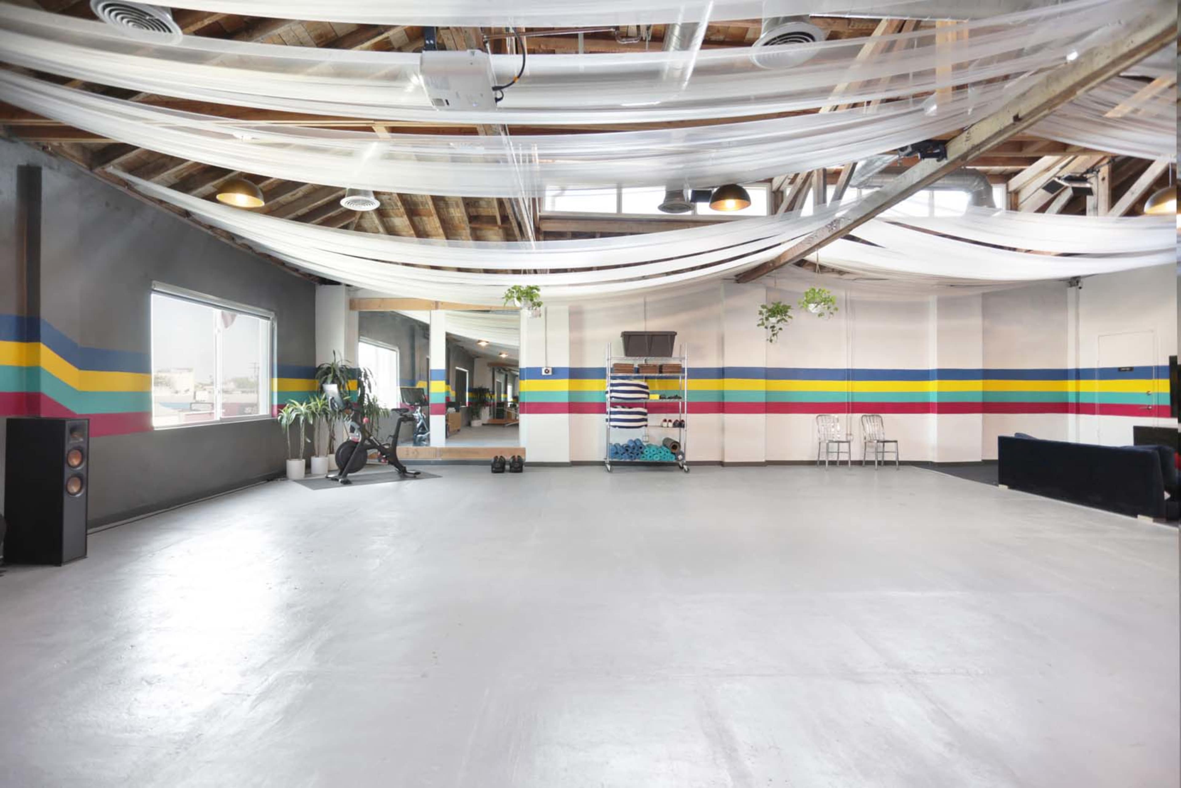 The image shows a spacious, industrial-style room with a polished floor, colorful wall stripes, and soft fabric draped from the ceiling.