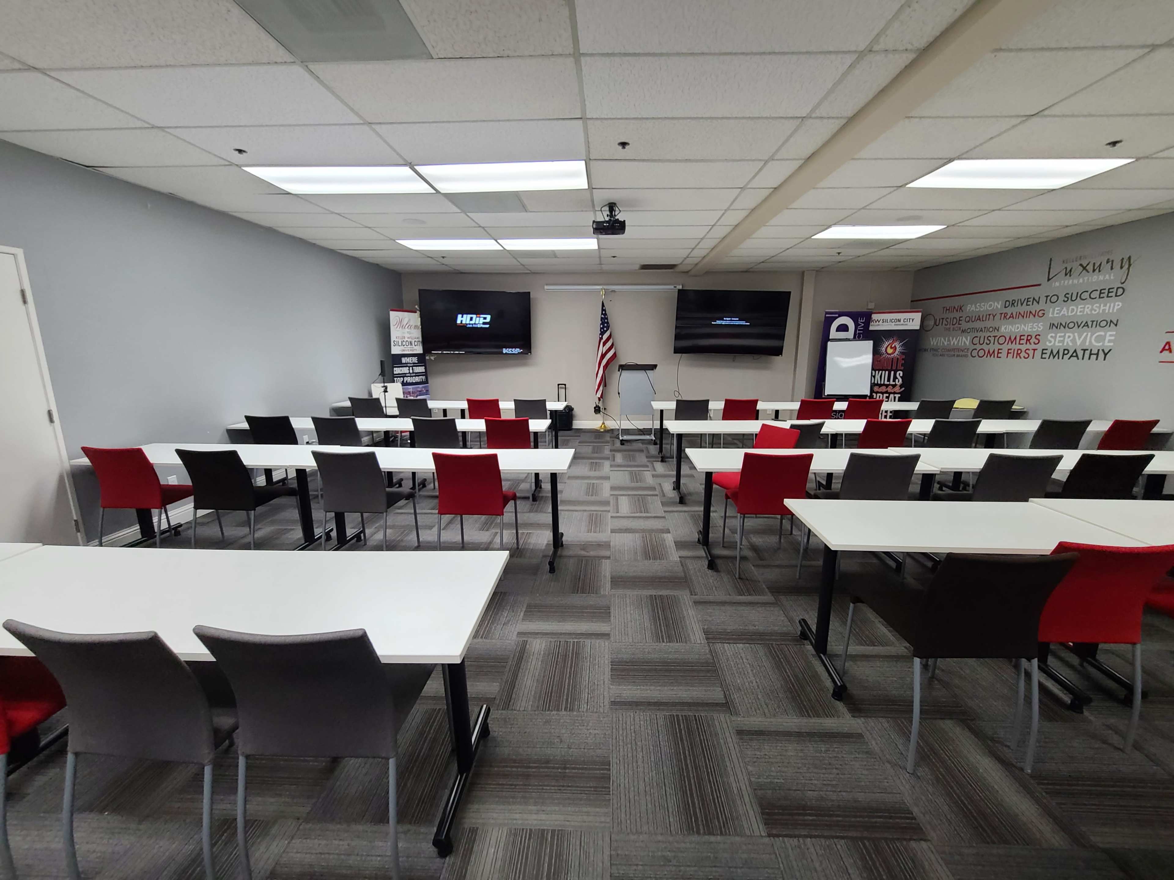 A classroom setup with several tables arranged in rows, an American flag, and screens mounted on the walls.
