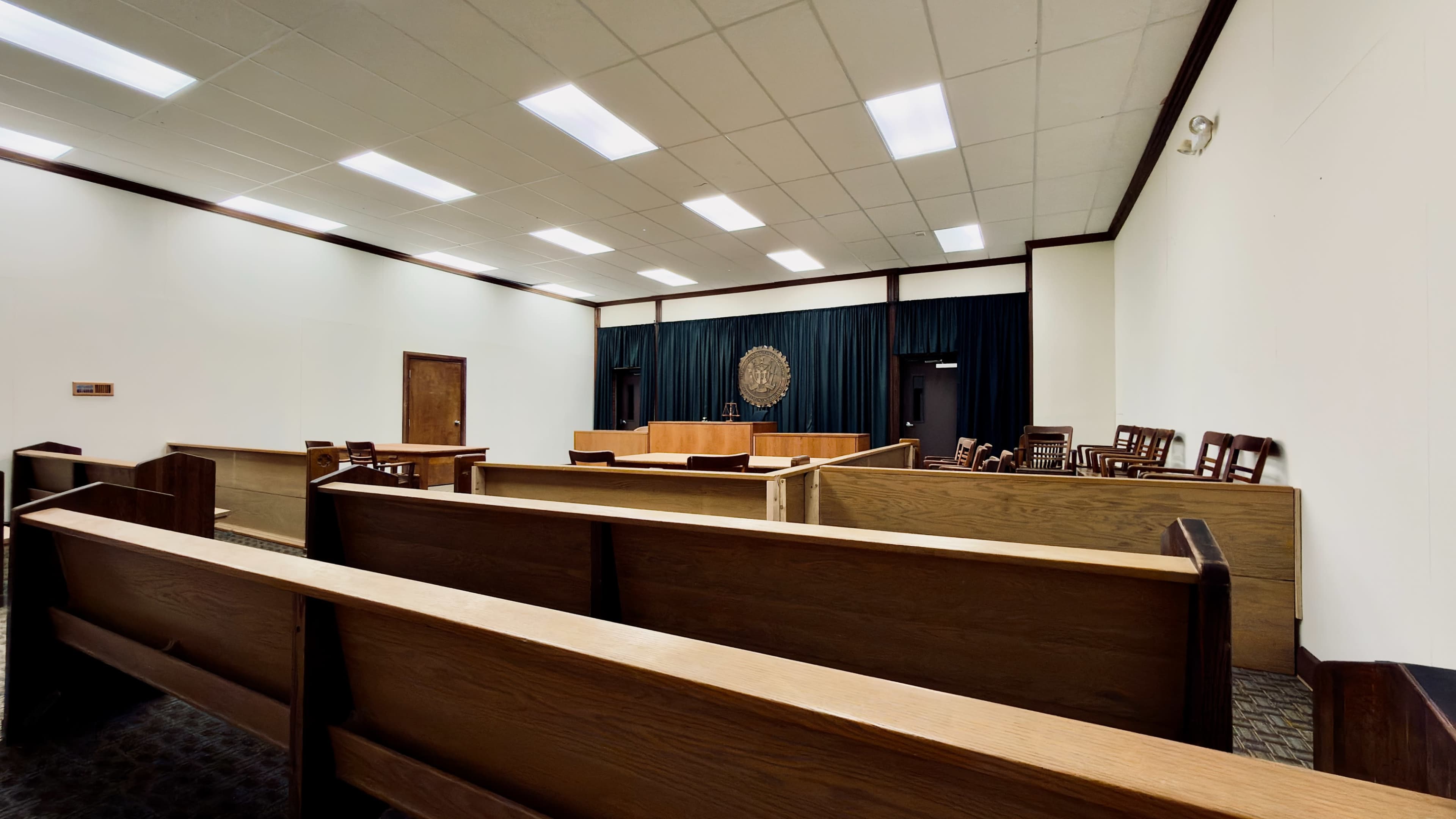 The image shows a sparsely furnished courtroom with wooden benches and a judge's bench at the front, flanked by a large emblem on the wall.