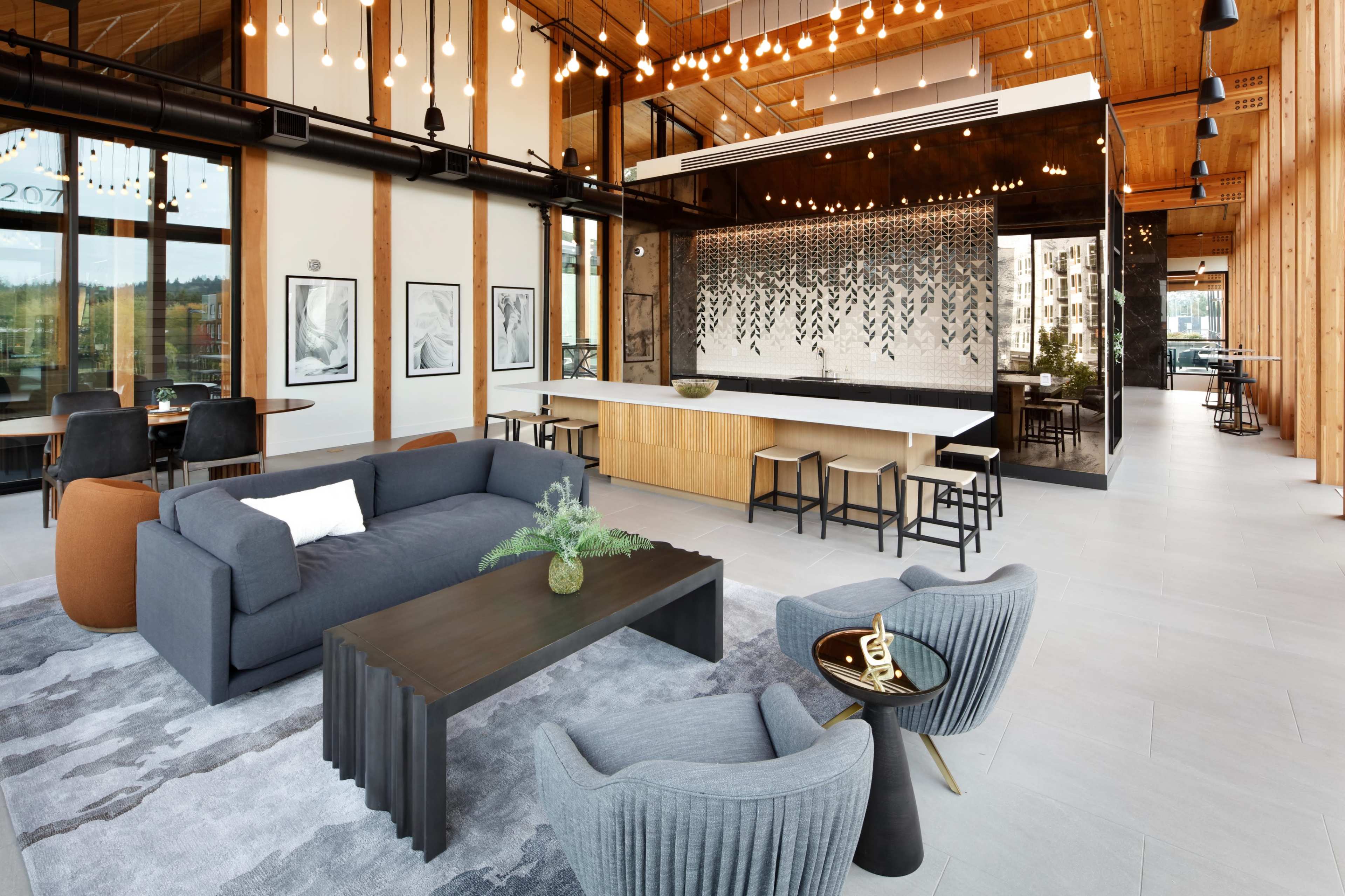 The image shows a modern, open-concept lounge area with wooden beams, stylish furniture, and a sleek kitchen bar under decorative lighting.