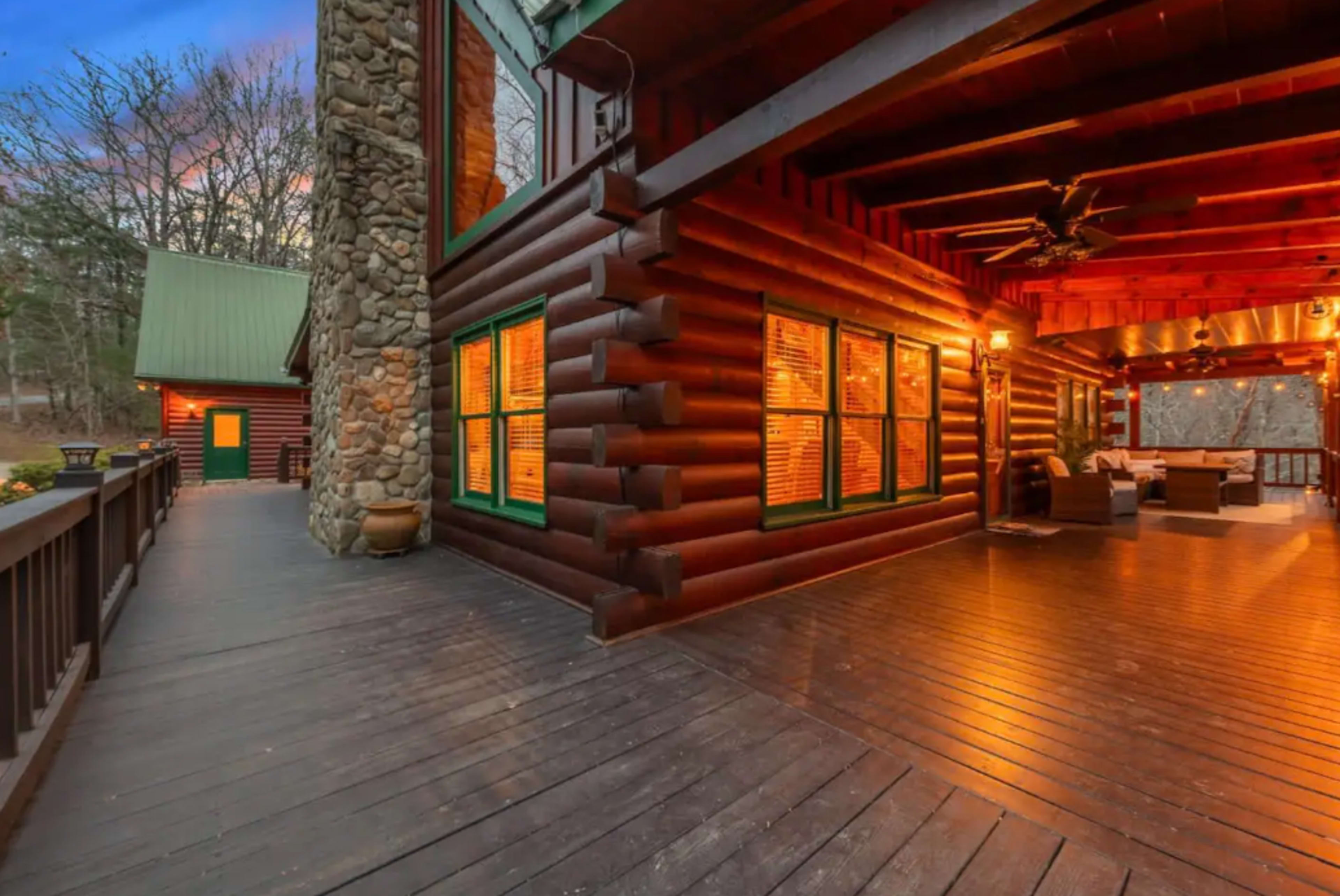 A wooden log cabin with a stone chimney, featuring illuminated windows and a spacious deck area.