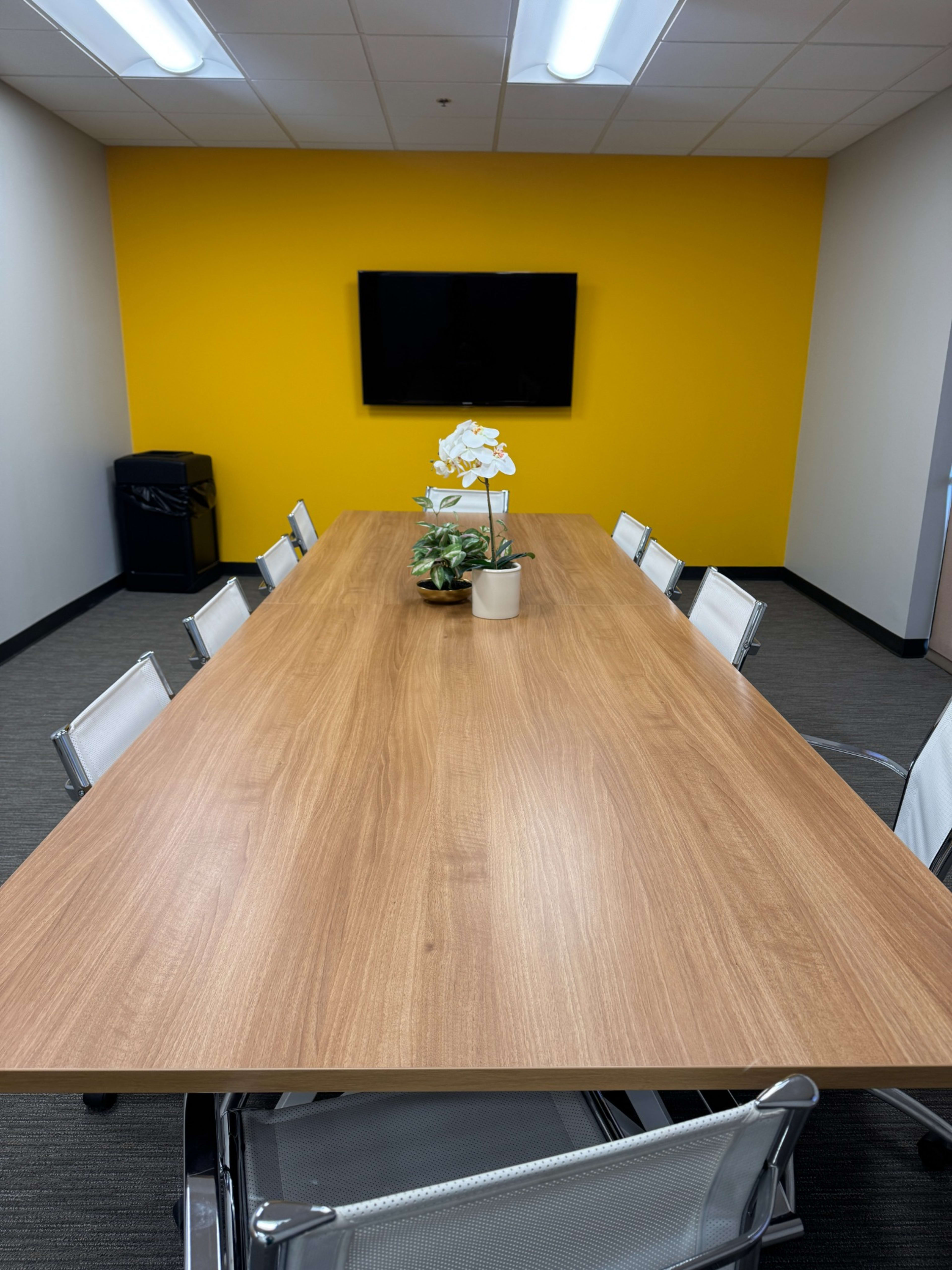 A long wooden conference table is set in a meeting room with a yellow accent wall and a wall-mounted television.