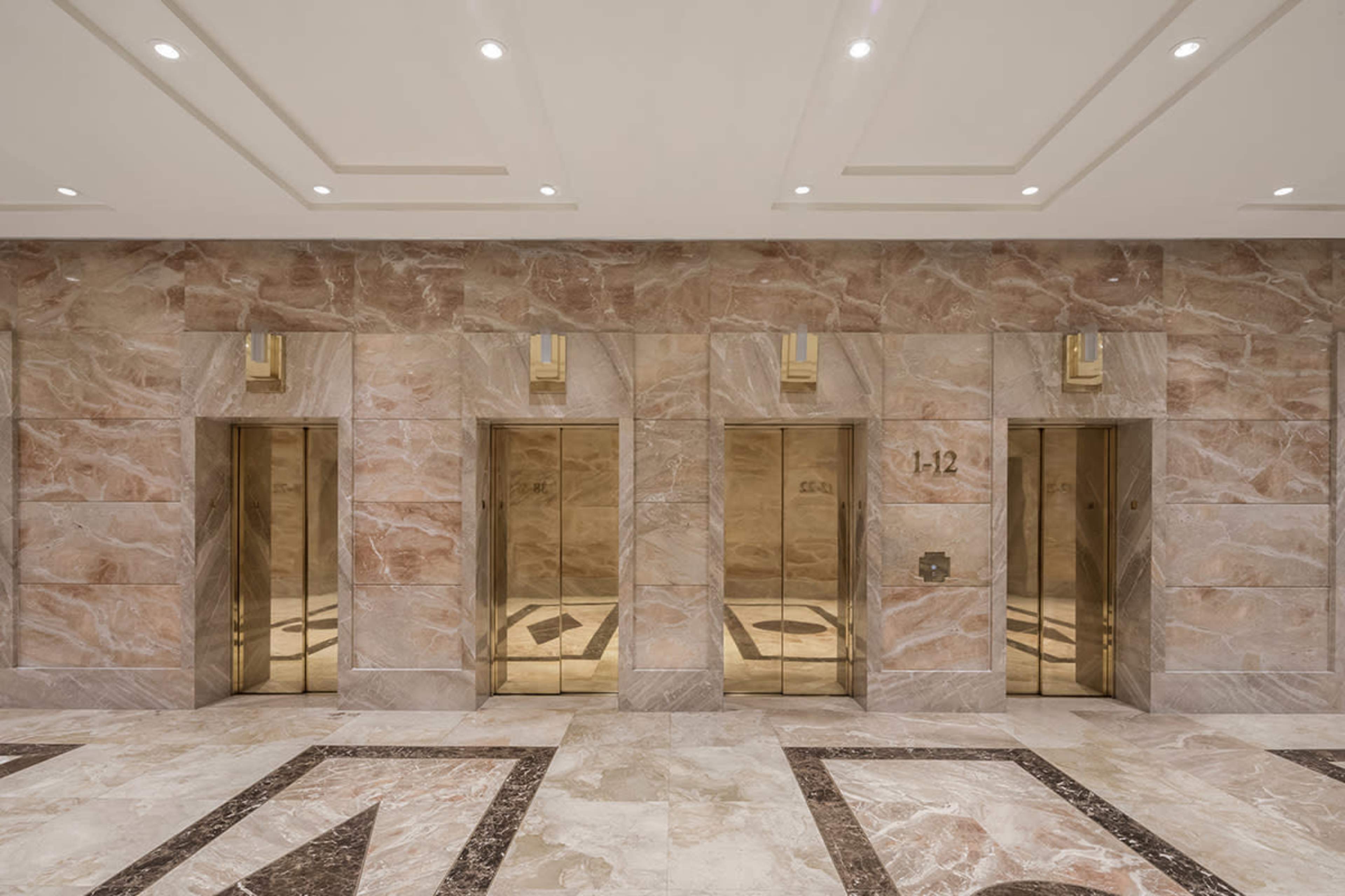 A spacious lobby with three elevator doors set in a wall of beige stone and marble flooring.