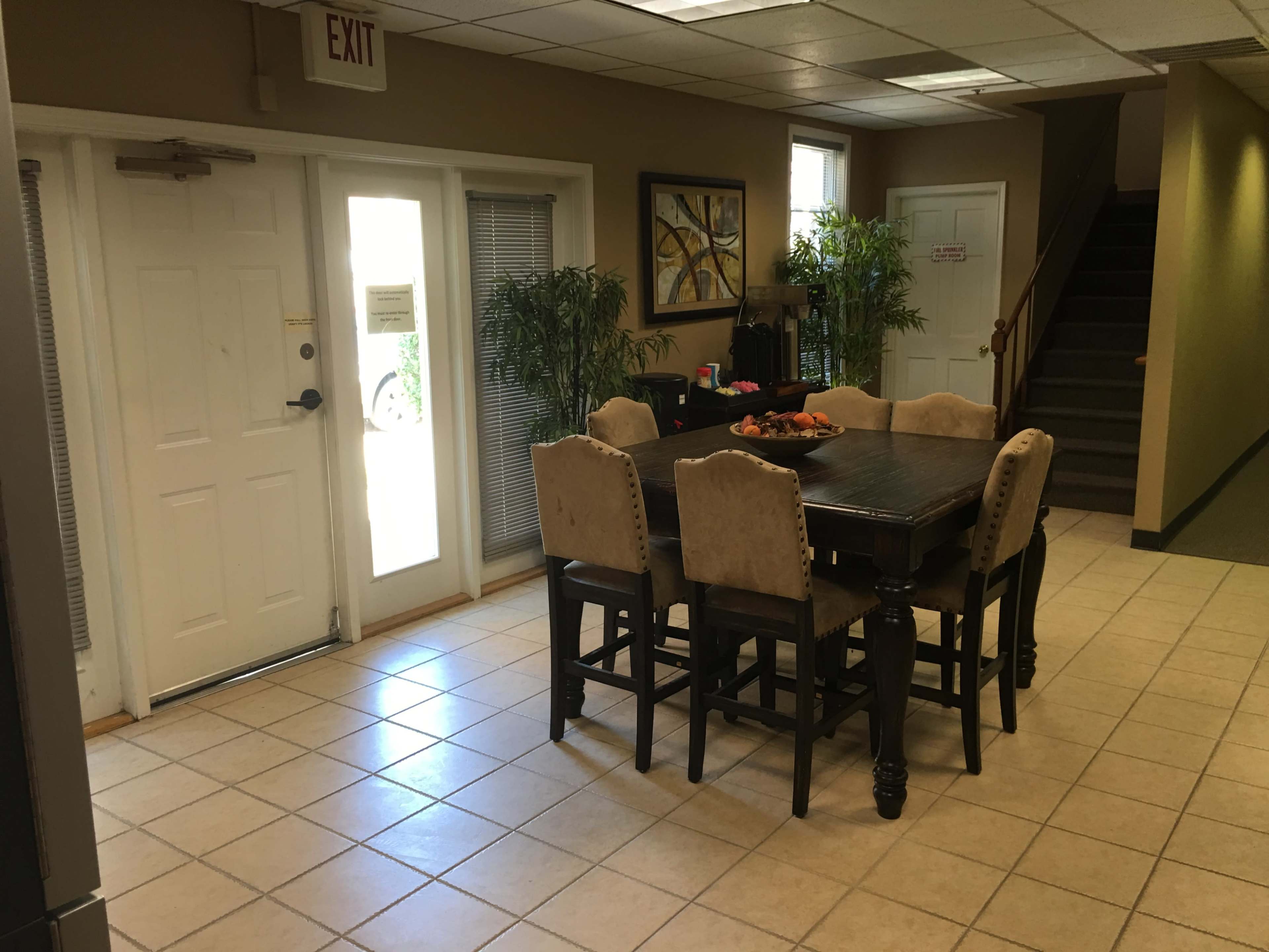 The image shows a well-lit room featuring a large wooden dining table surrounded by upholstered chairs, with an exit door and a staircase visible in the background.