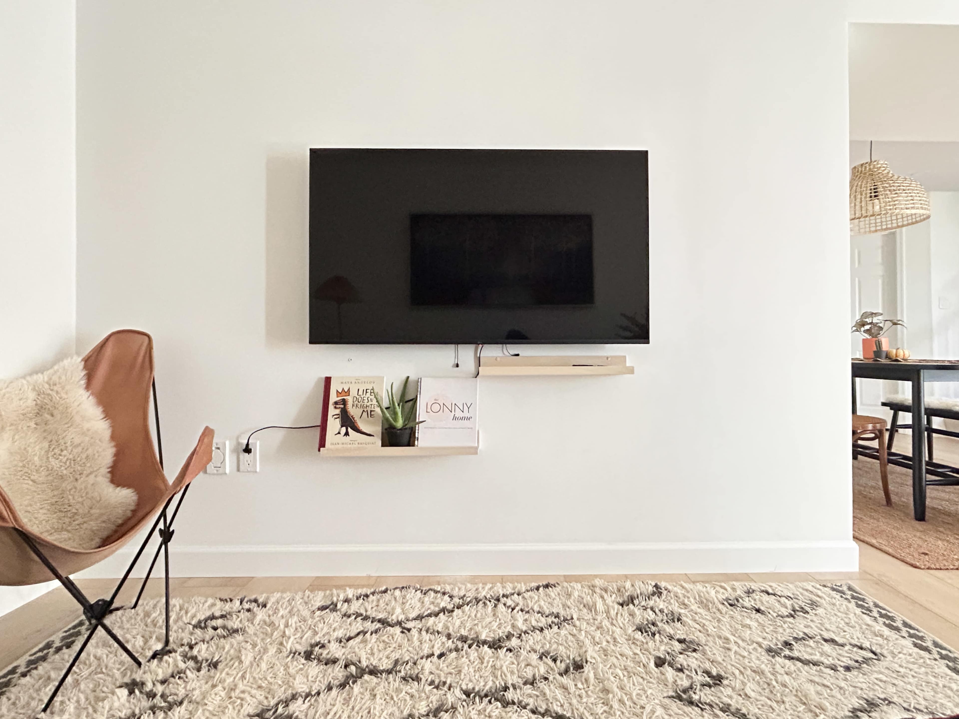 A wall-mounted television is positioned above a small shelf with two books, accompanied by a butterfly chair on a patterned rug in a minimalist room.