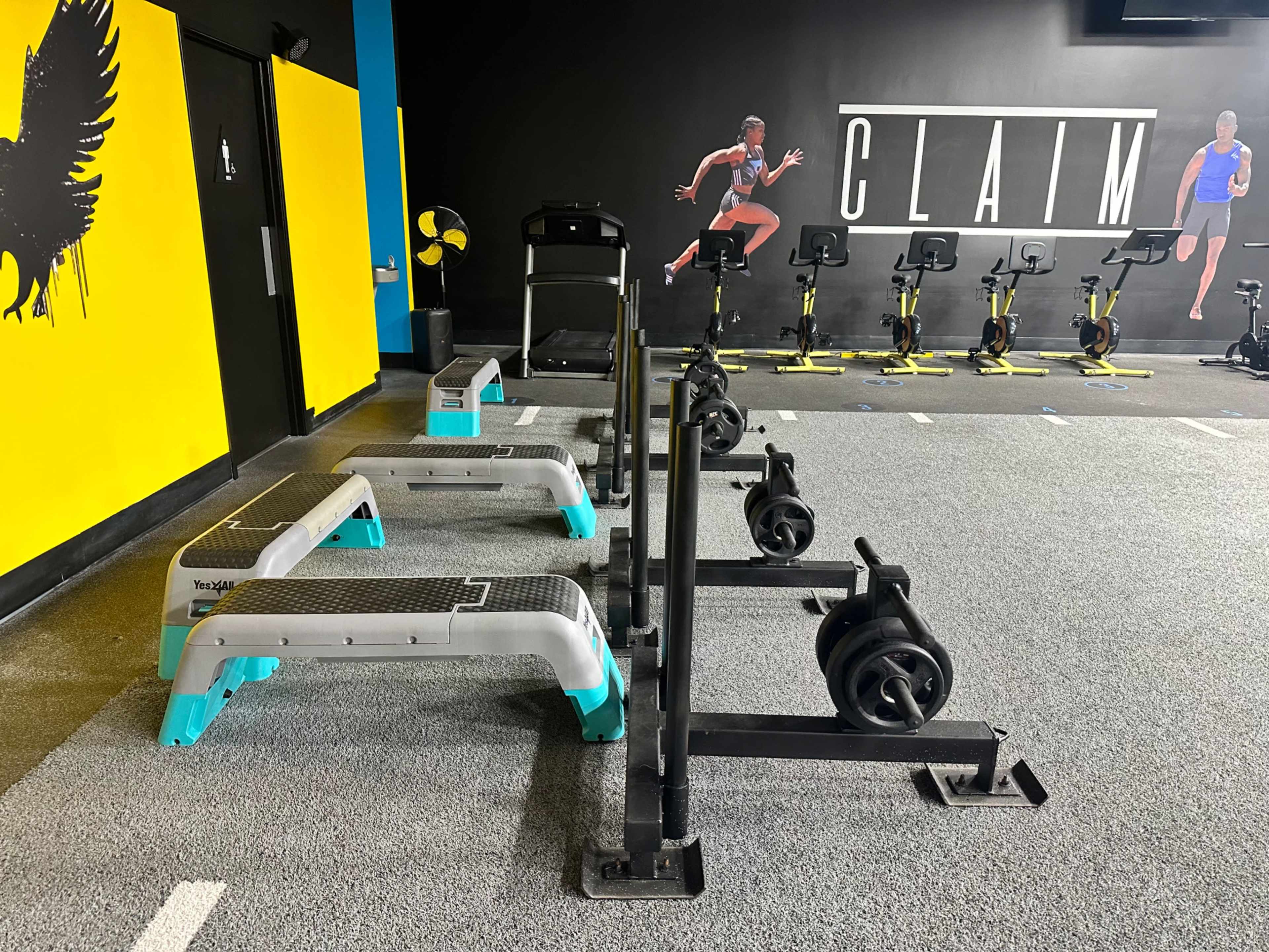 A fitness area with exercise equipment, including step platforms and a treadmill, against a black wall featuring athletic mural art.