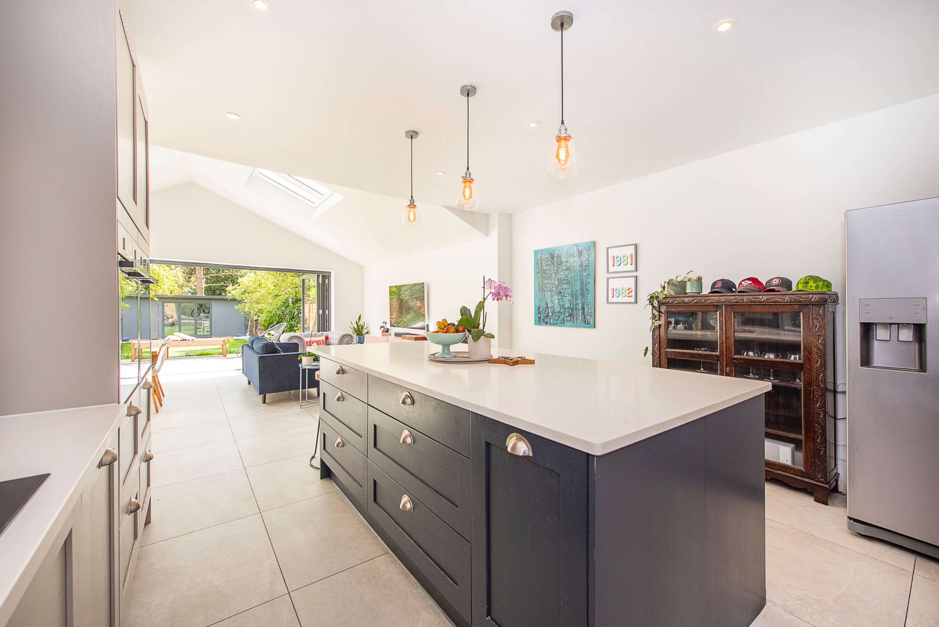 The image shows a modern kitchen with a large island, pendant lighting, and an open layout leading to a garden view.
