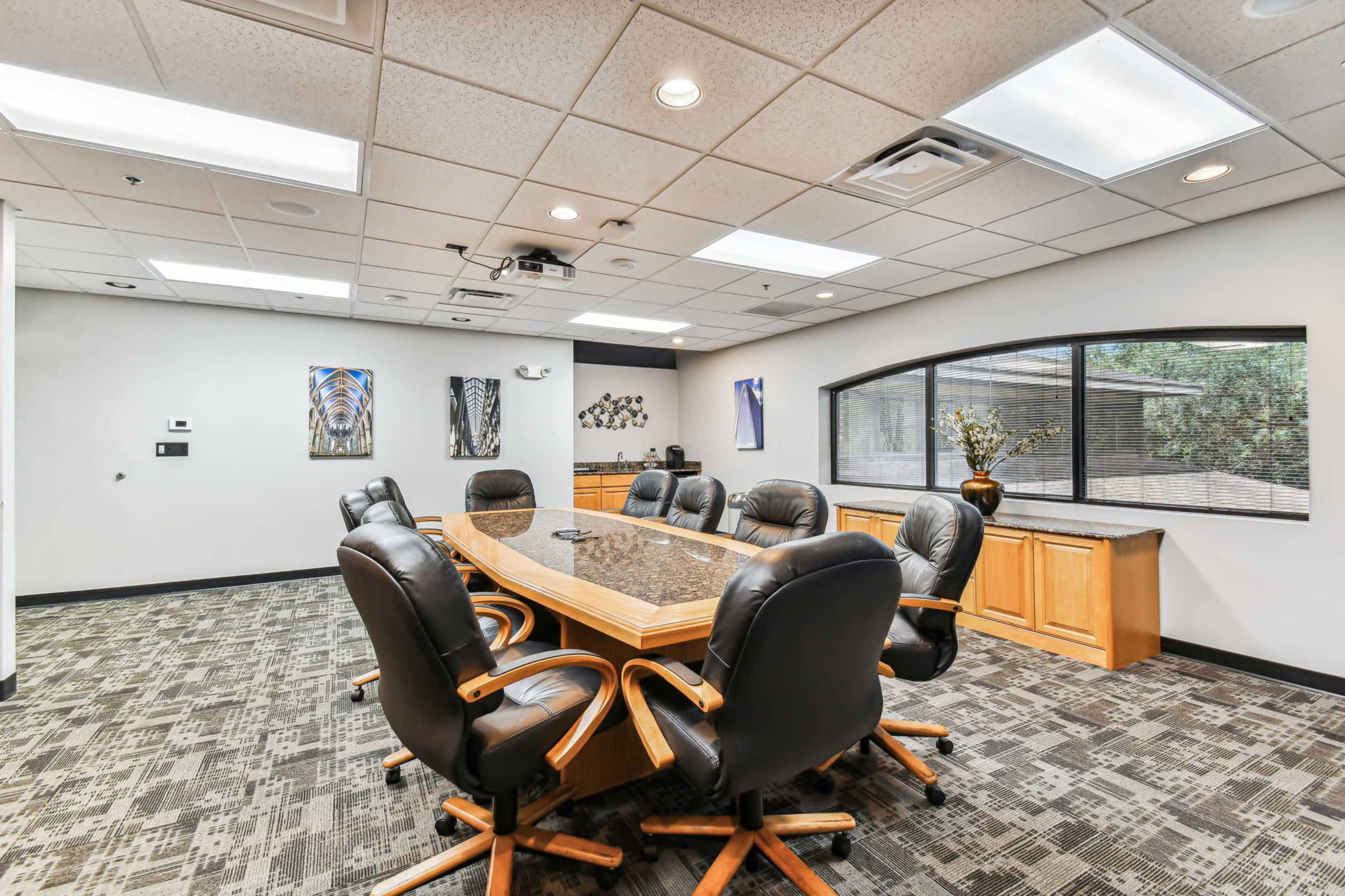 The image shows a modern conference room with a rectangular table surrounded by ergonomic black chairs and equipped with a projector and large windows.
