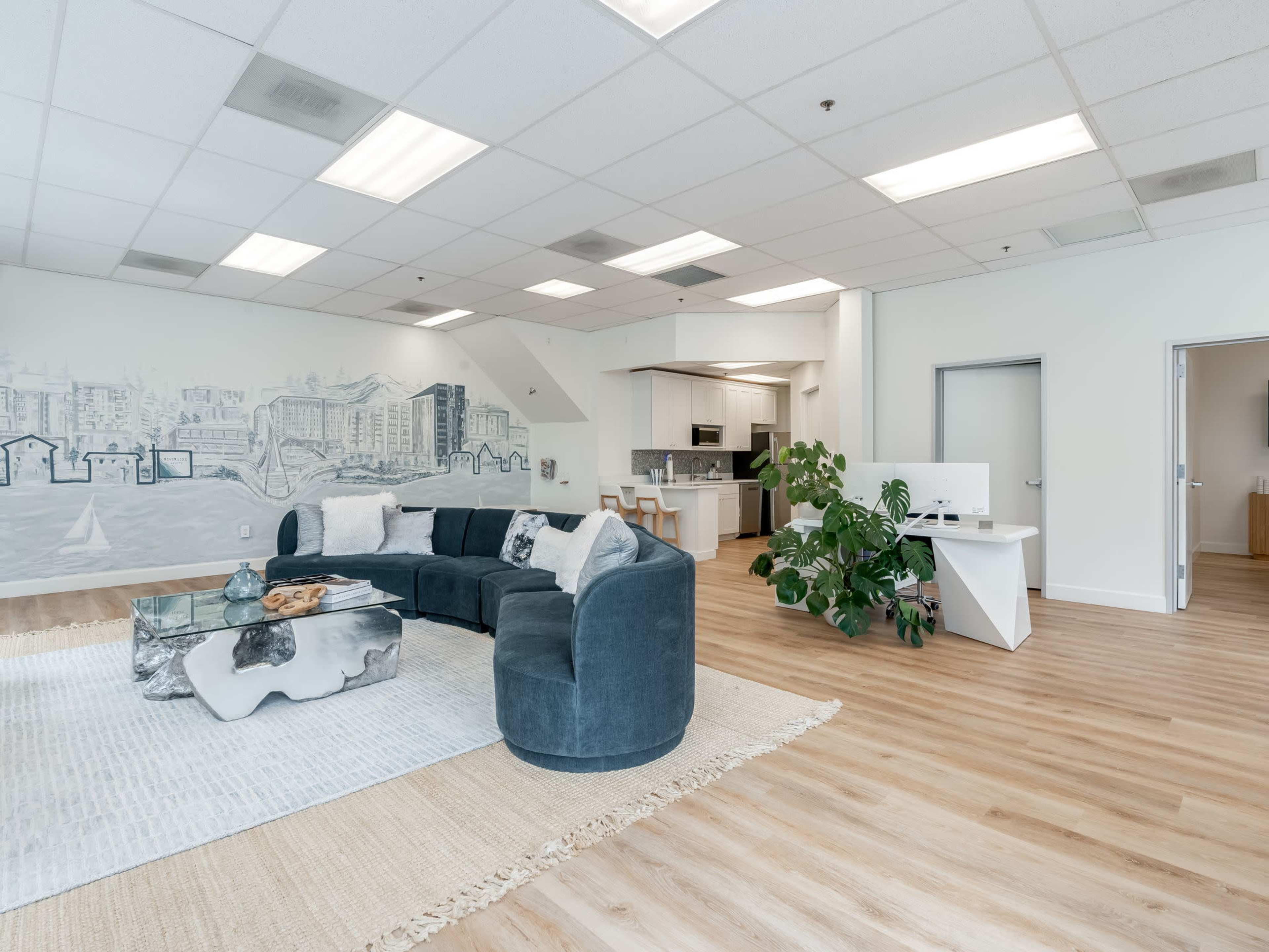 The open-concept space features a curved sofa, a glass coffee table, and a view of a kitchen area, with a mural depicting a cityscape on one wall.