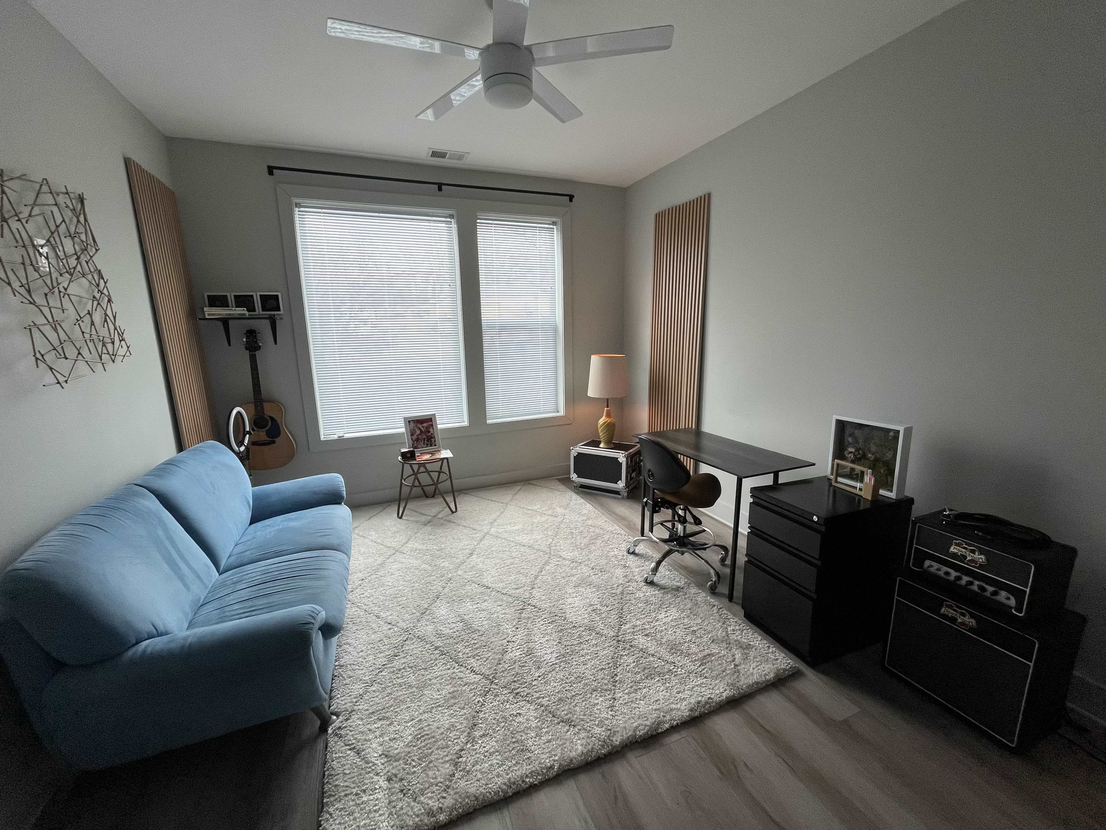 The image depicts a modern living room with a blue couch, a desk and chair, a lamp, and a record player, complemented by large windows and light-colored walls.