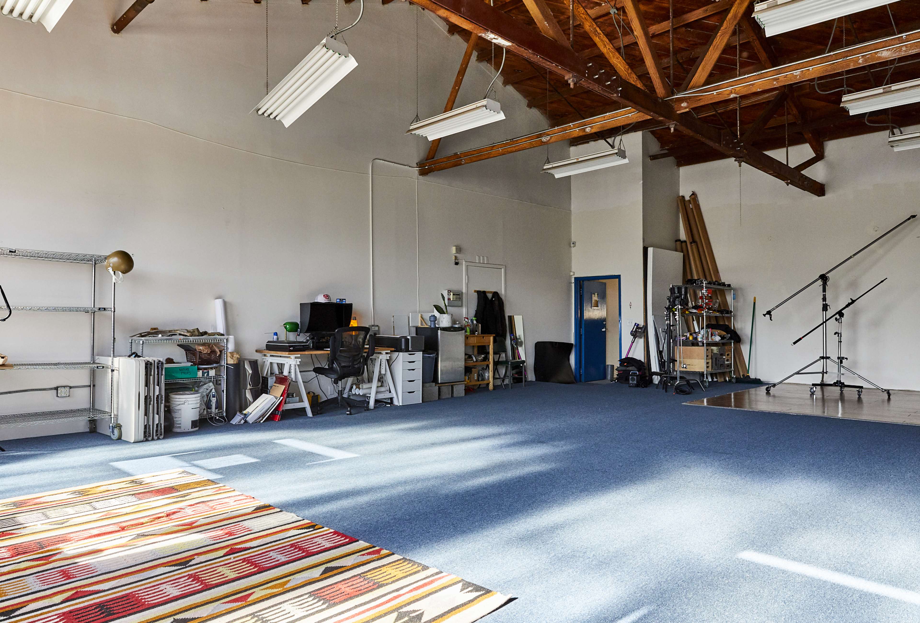 An empty studio space features wooden beams, blue carpet, and workstations along one side, with various equipment and tools scattered throughout.
