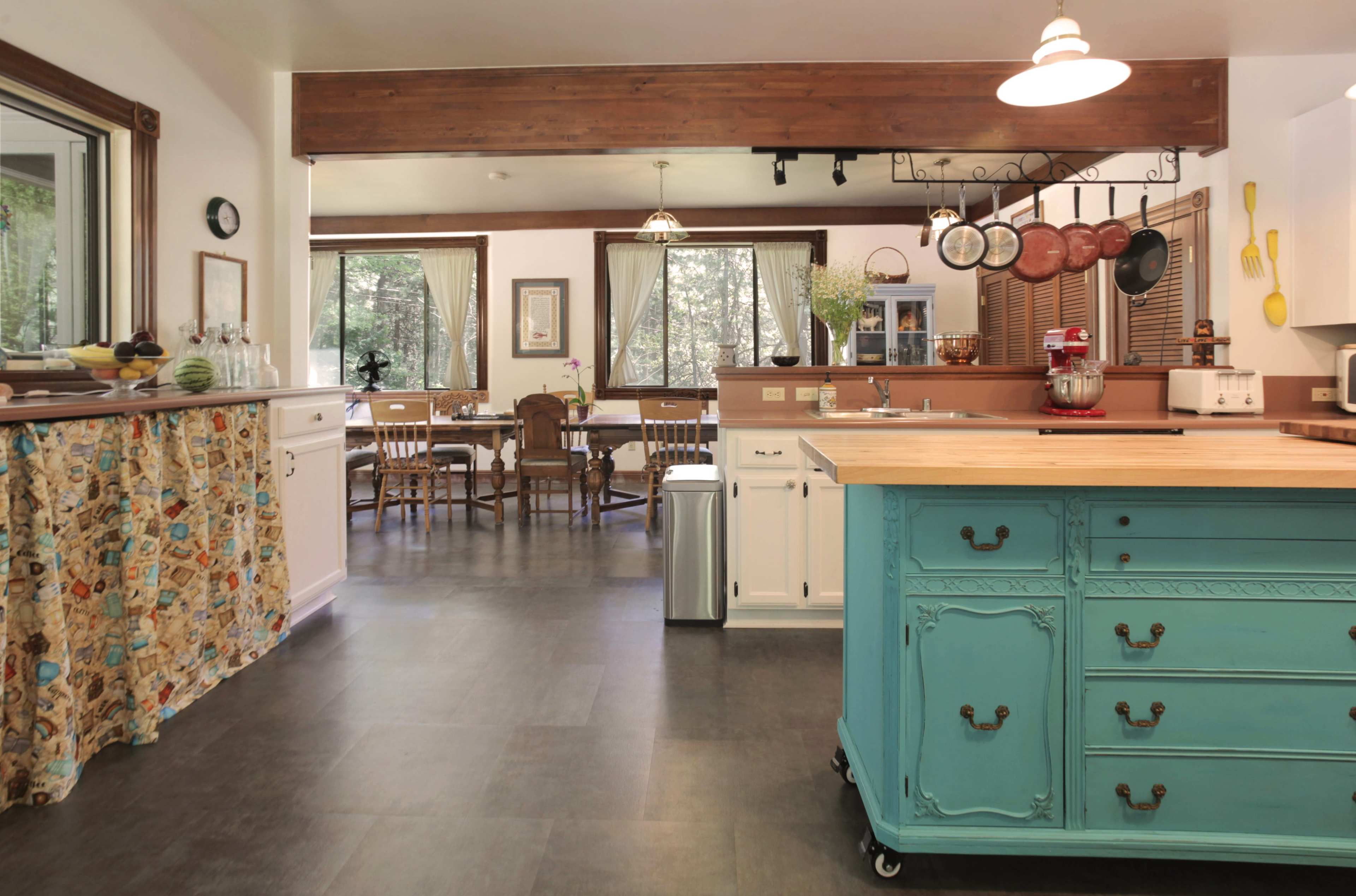 A spacious kitchen features a central island with a turquoise base, white cabinets, and a dining area in the background surrounded by windows.