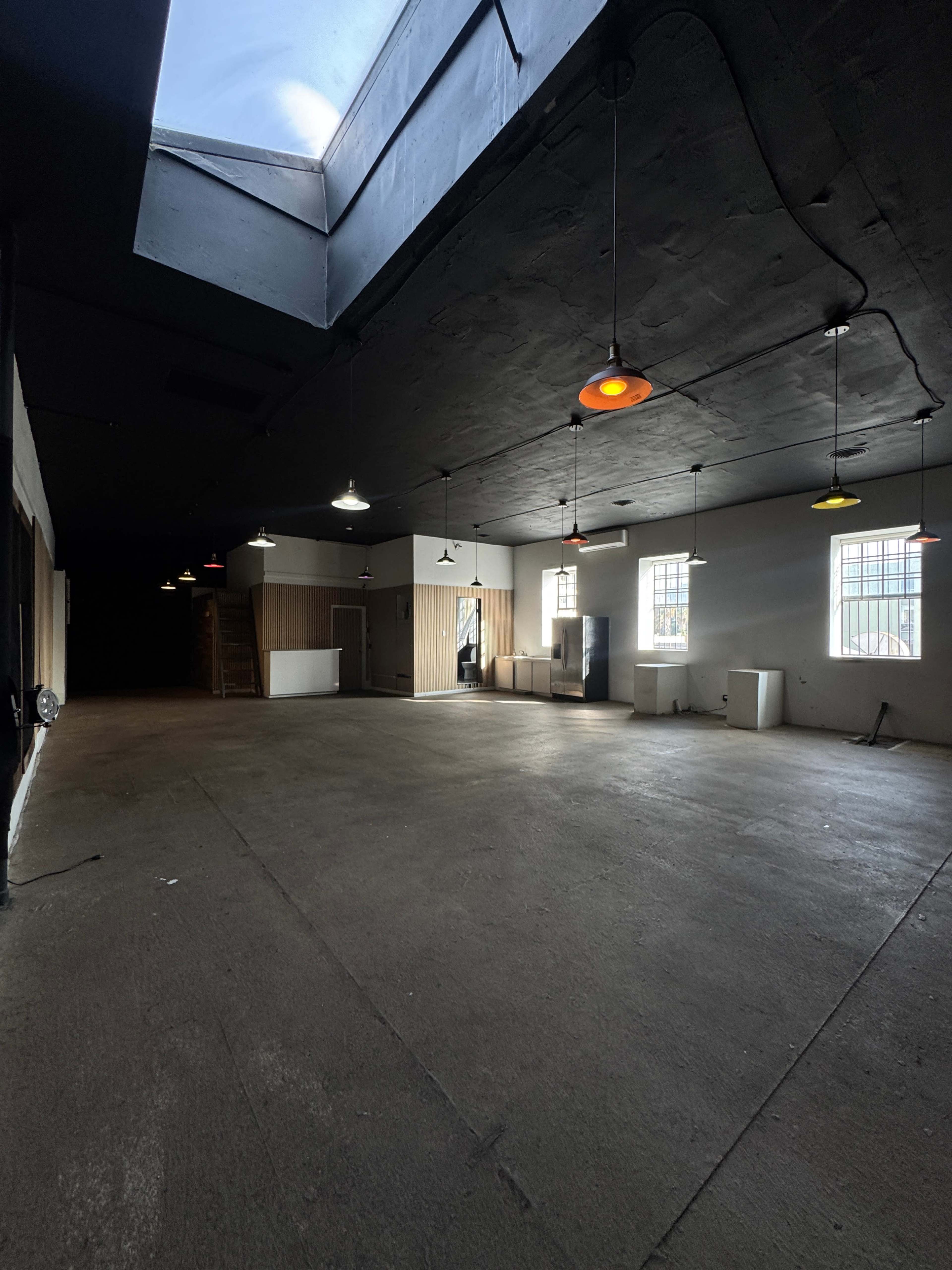The image shows a large, empty interior space with concrete flooring, illuminated by pendant lights and featuring a skylight.