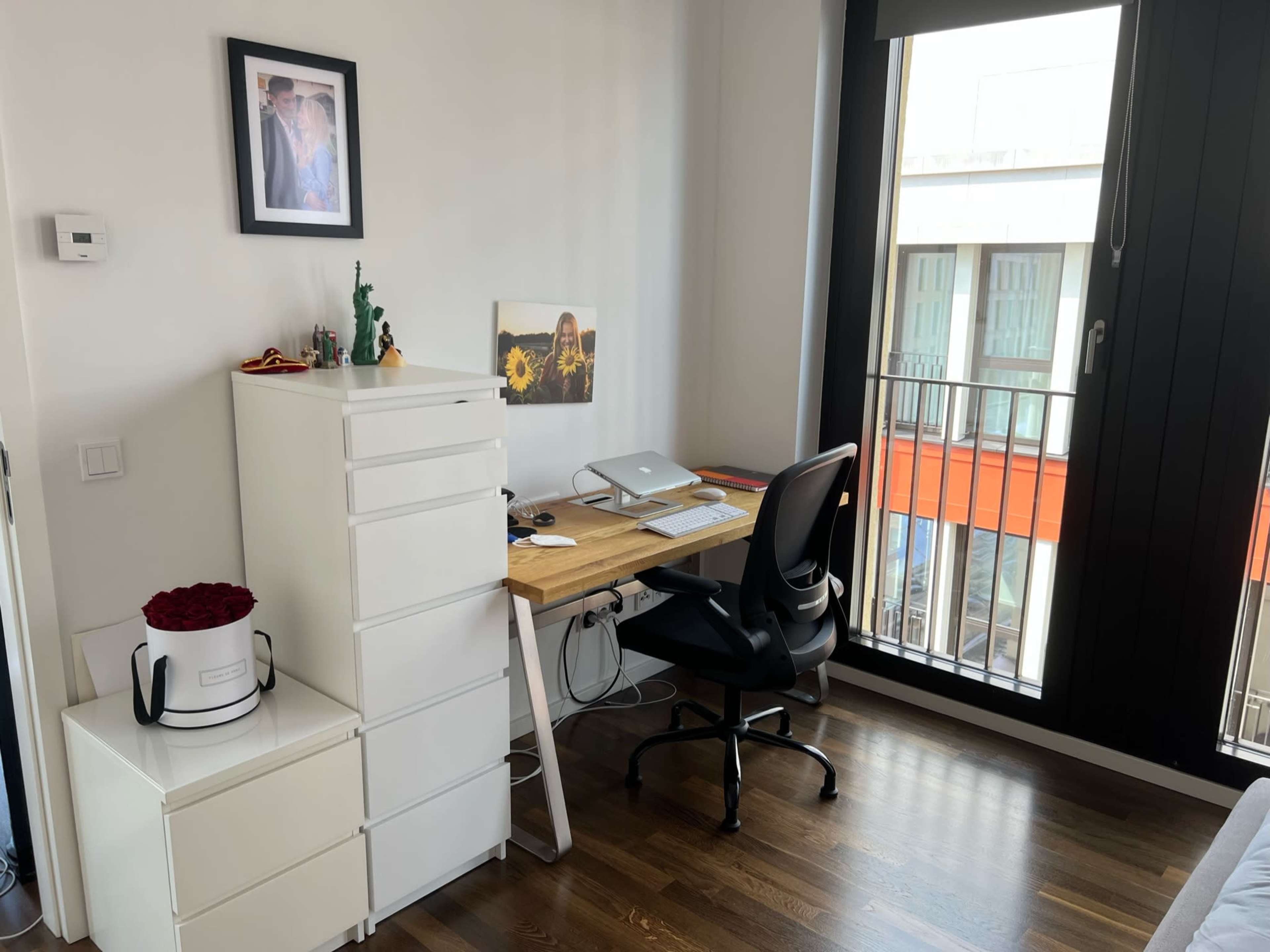 A modern workspace featuring a desk with a computer, a black chair, a white storage unit, and a window overlooking a balcony.