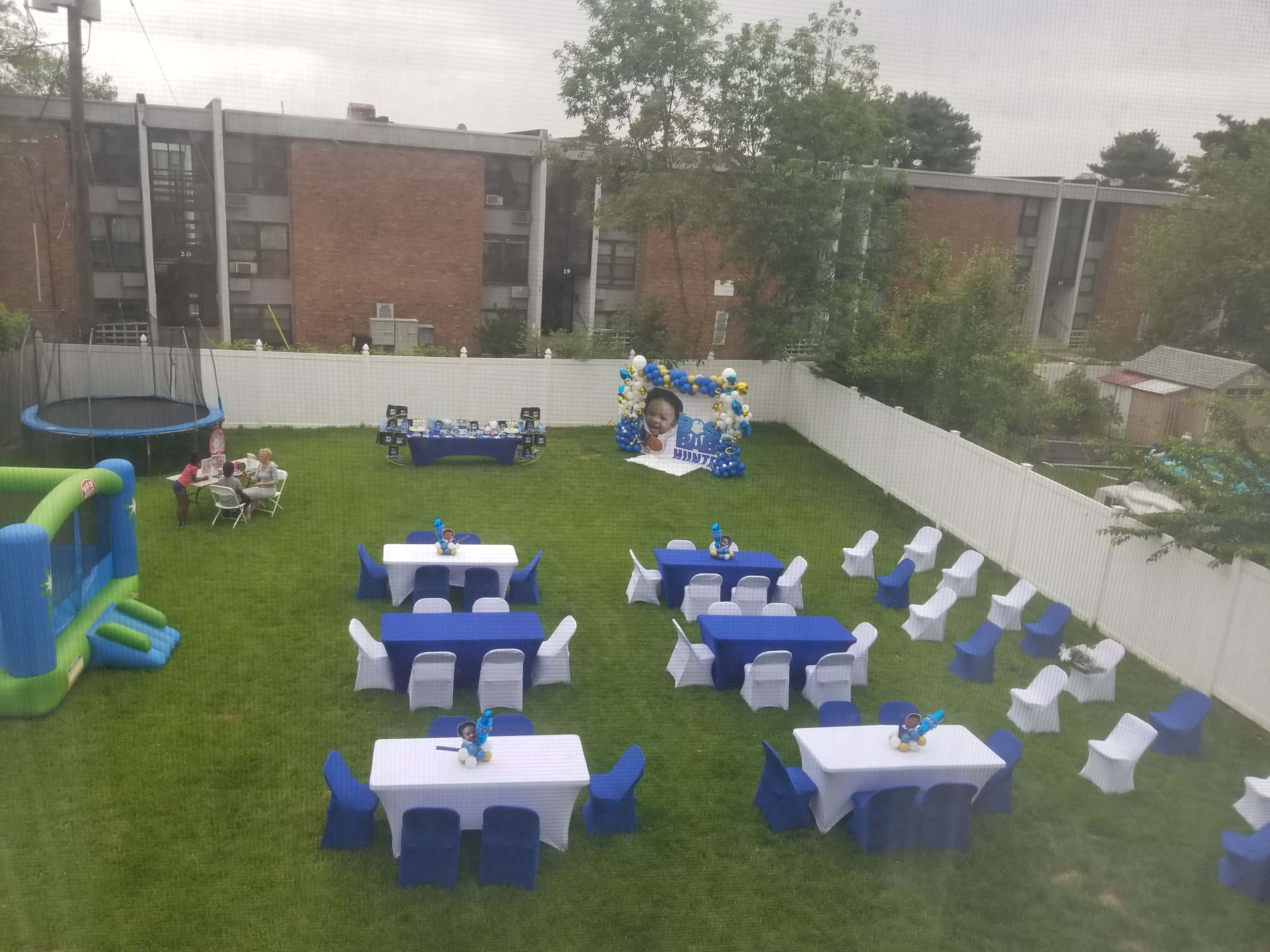A backyard is set up for a party with white tables covered in blue cloth, decorated with centerpieces, and a large balloon display in the background.