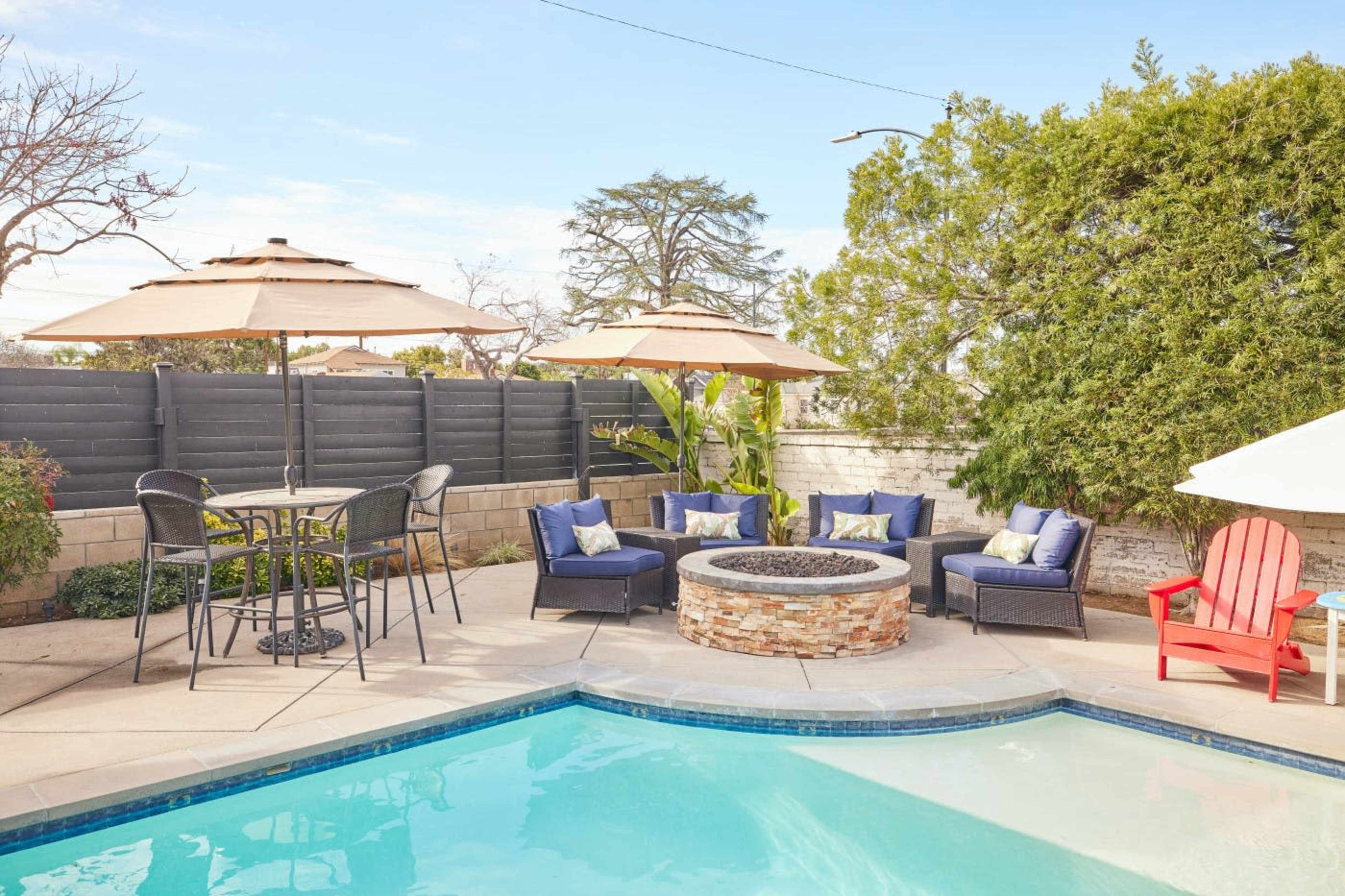 The image shows a backyard with a swimming pool, a stone fire pit surrounded by seating, and patio umbrellas providing shade.