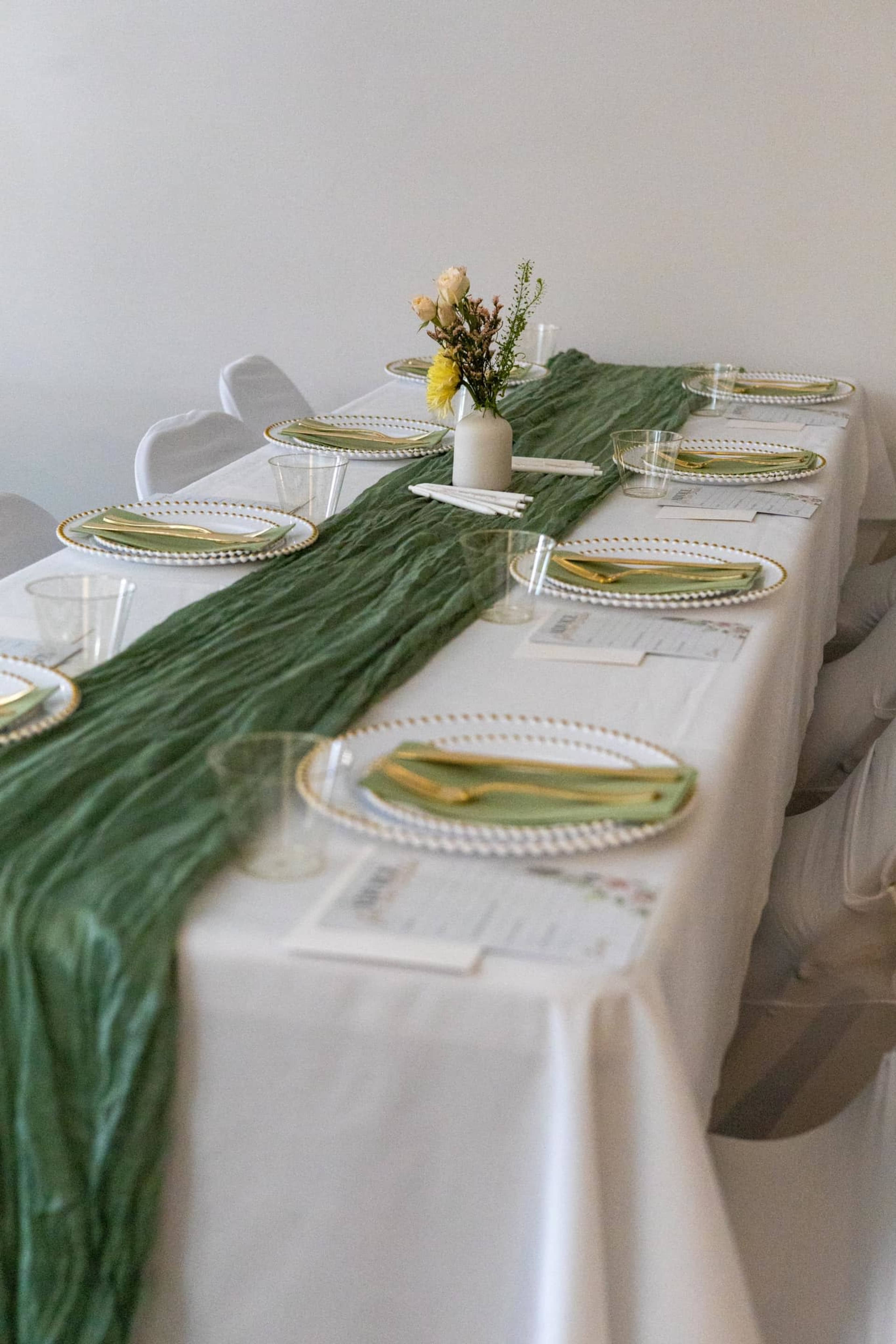 A long, elegantly set dining table features a green runner, floral centerpiece, plates, and glassware arranged for multiple guests.