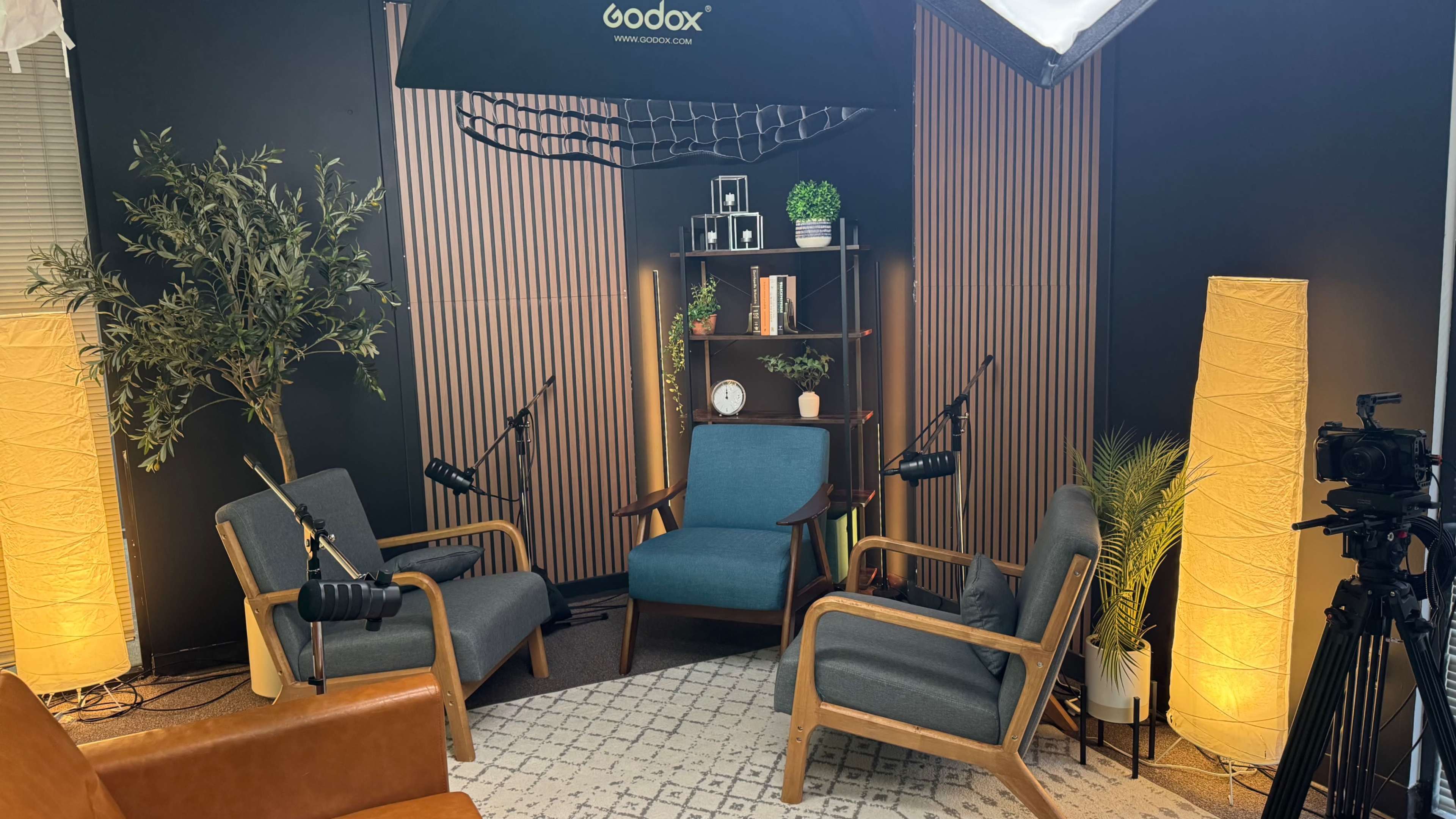 The image shows a modern studio setup with three chairs, a bookshelf, and soft lighting, creating a relaxed atmosphere for interviews or discussions.