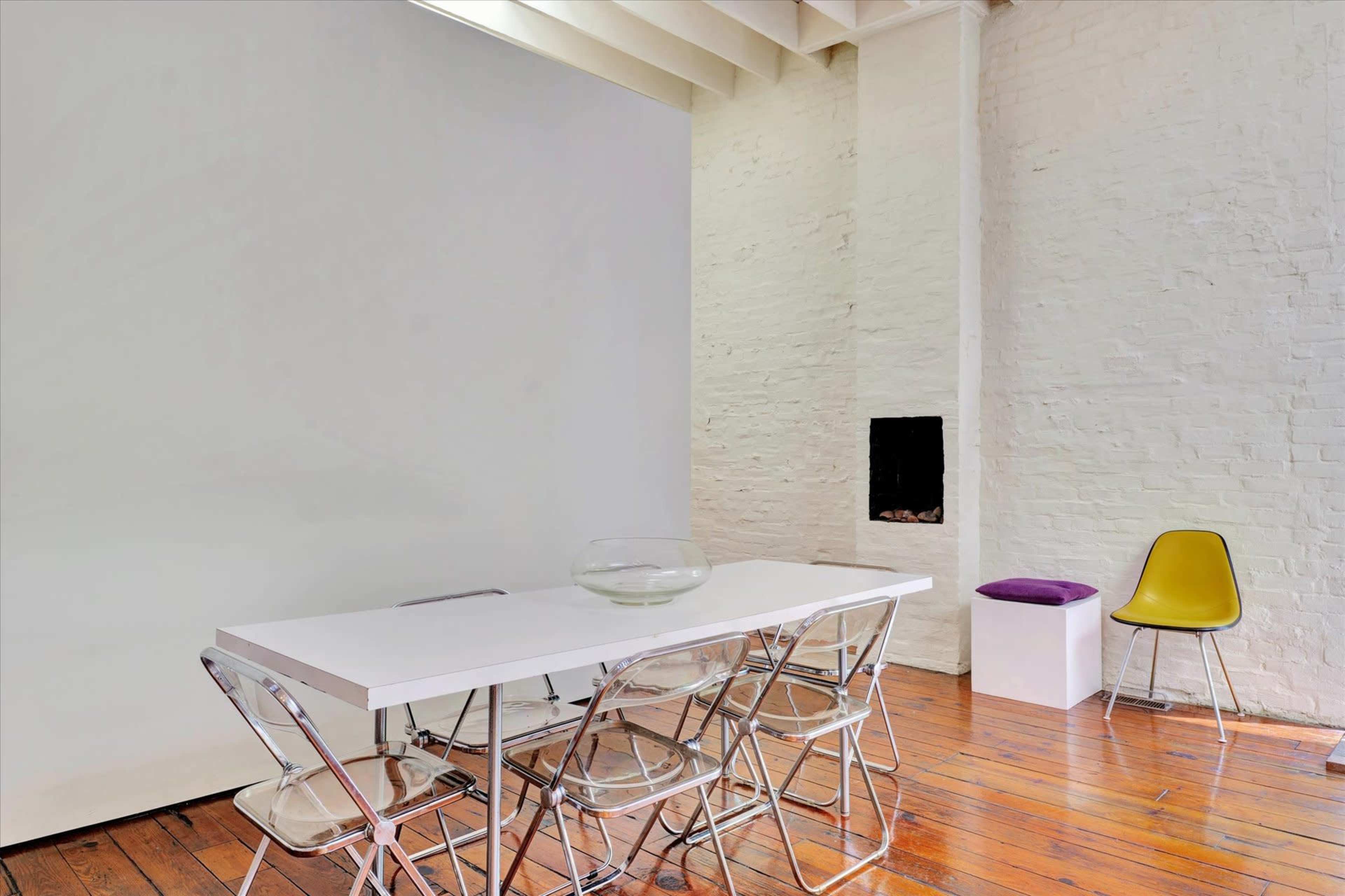 A minimalist dining area features a white table with silver folding chairs, a small decorative bowl, and a single yellow chair beside a white wall.