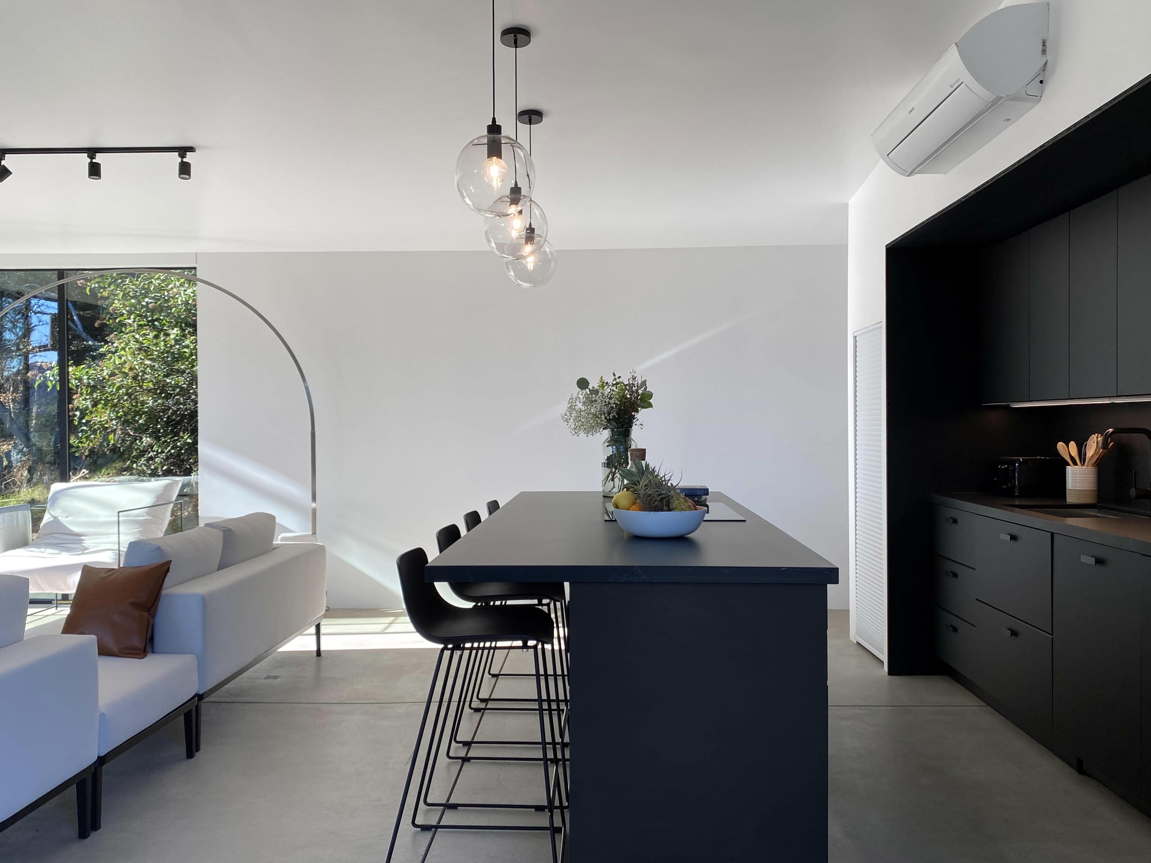 A modern kitchen with a black island bar, minimalist decor, and large windows allowing natural light to enter the space.