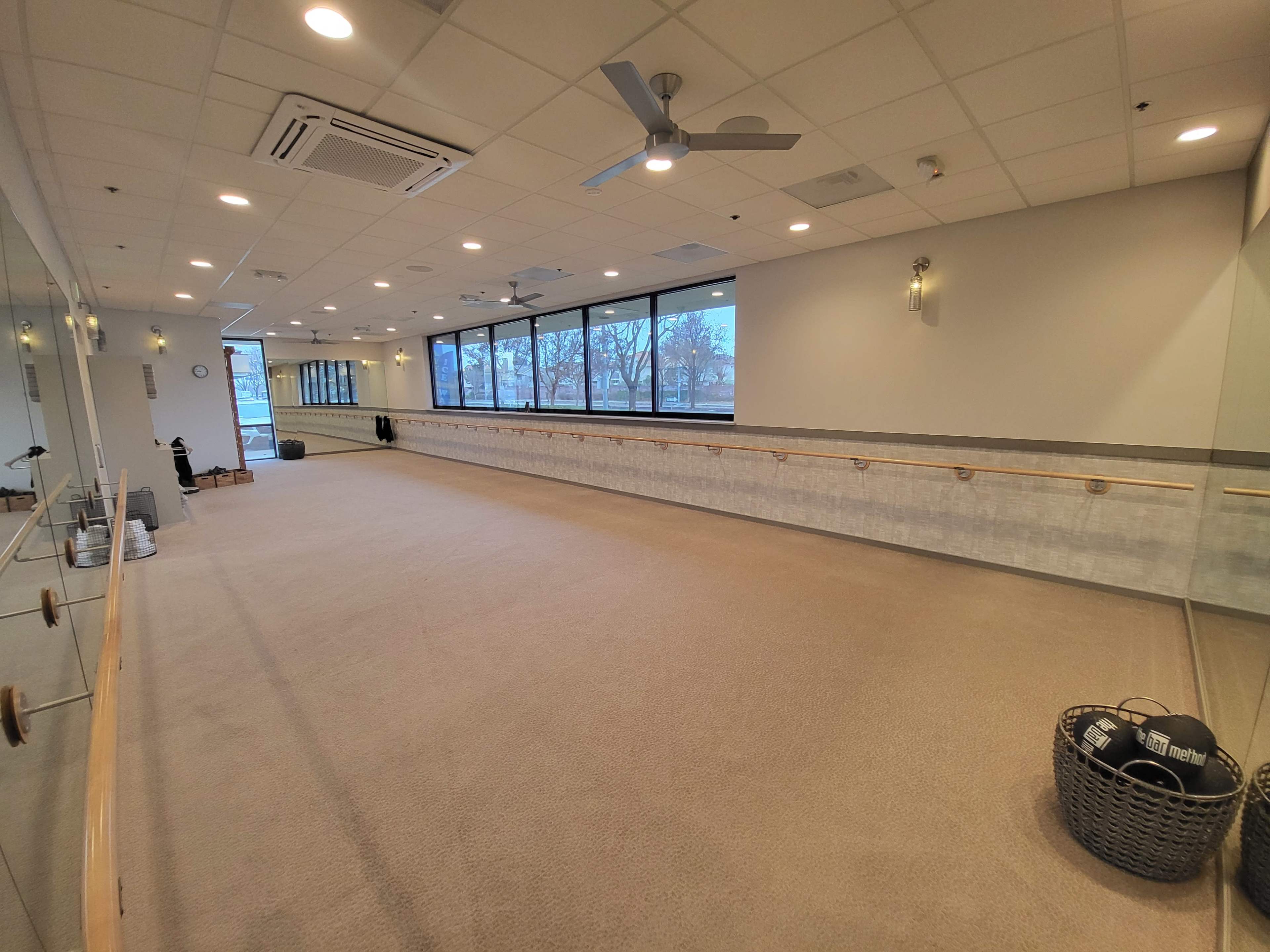 A spacious, well-lit exercise studio with a mirrored wall and a ballet bar, featuring large windows that provide natural light.