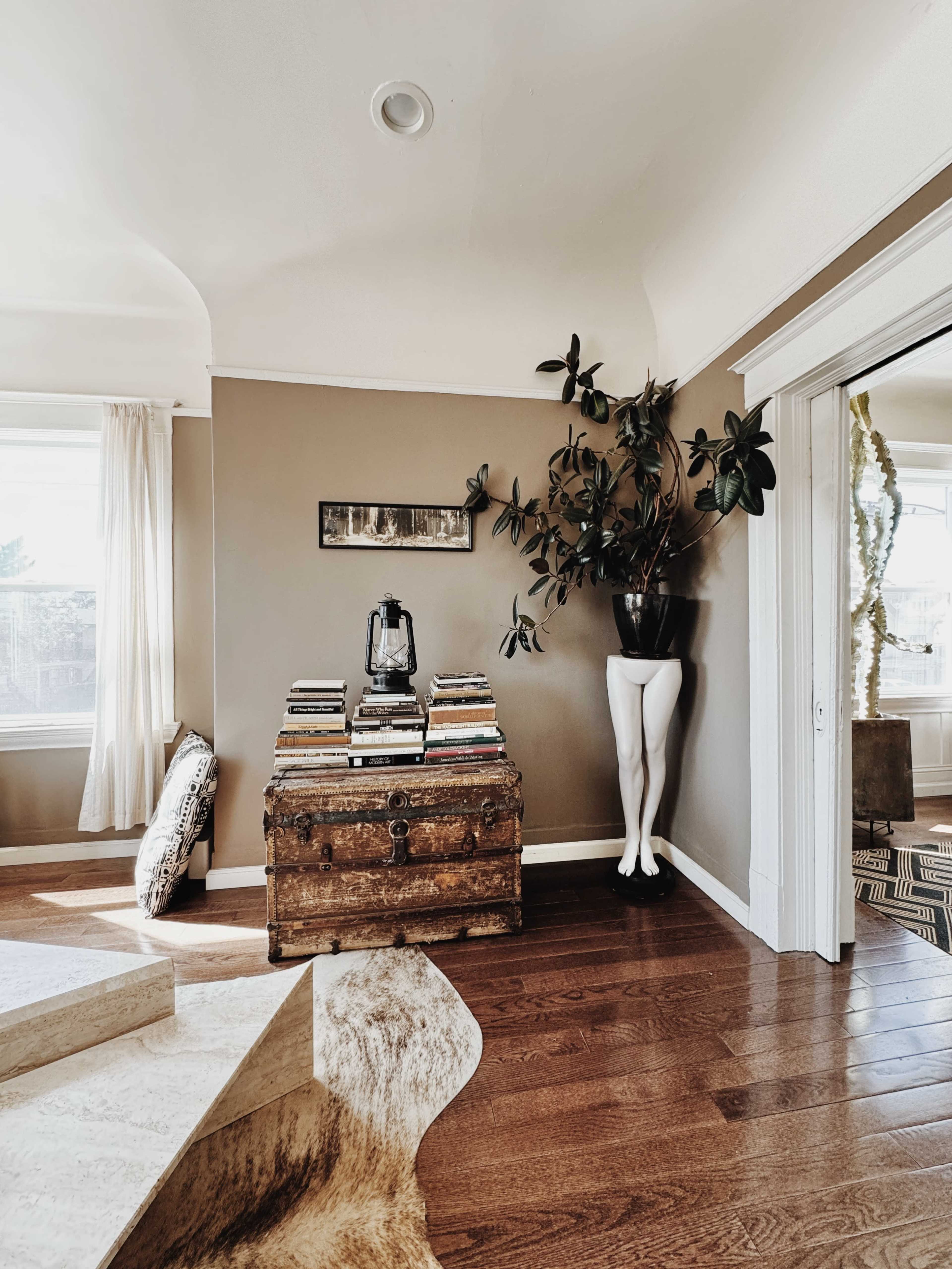 The image shows a cozy room with a wooden chest, stacked books, a potted plant, and a unique planter resembling a pair of legs in the corner.