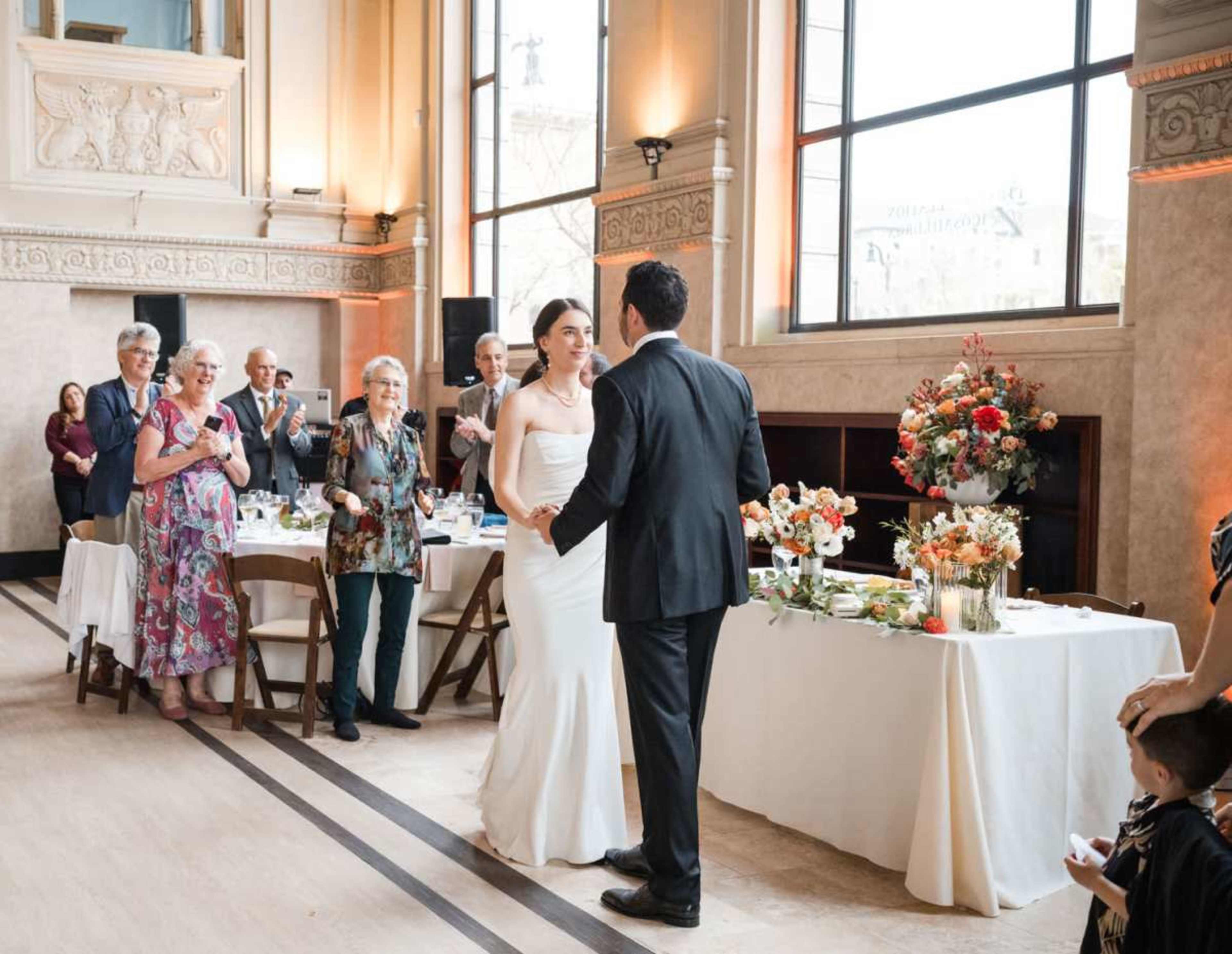 A bride and groom stand together in a decorated hall, while guests applaud and witness their celebration.
