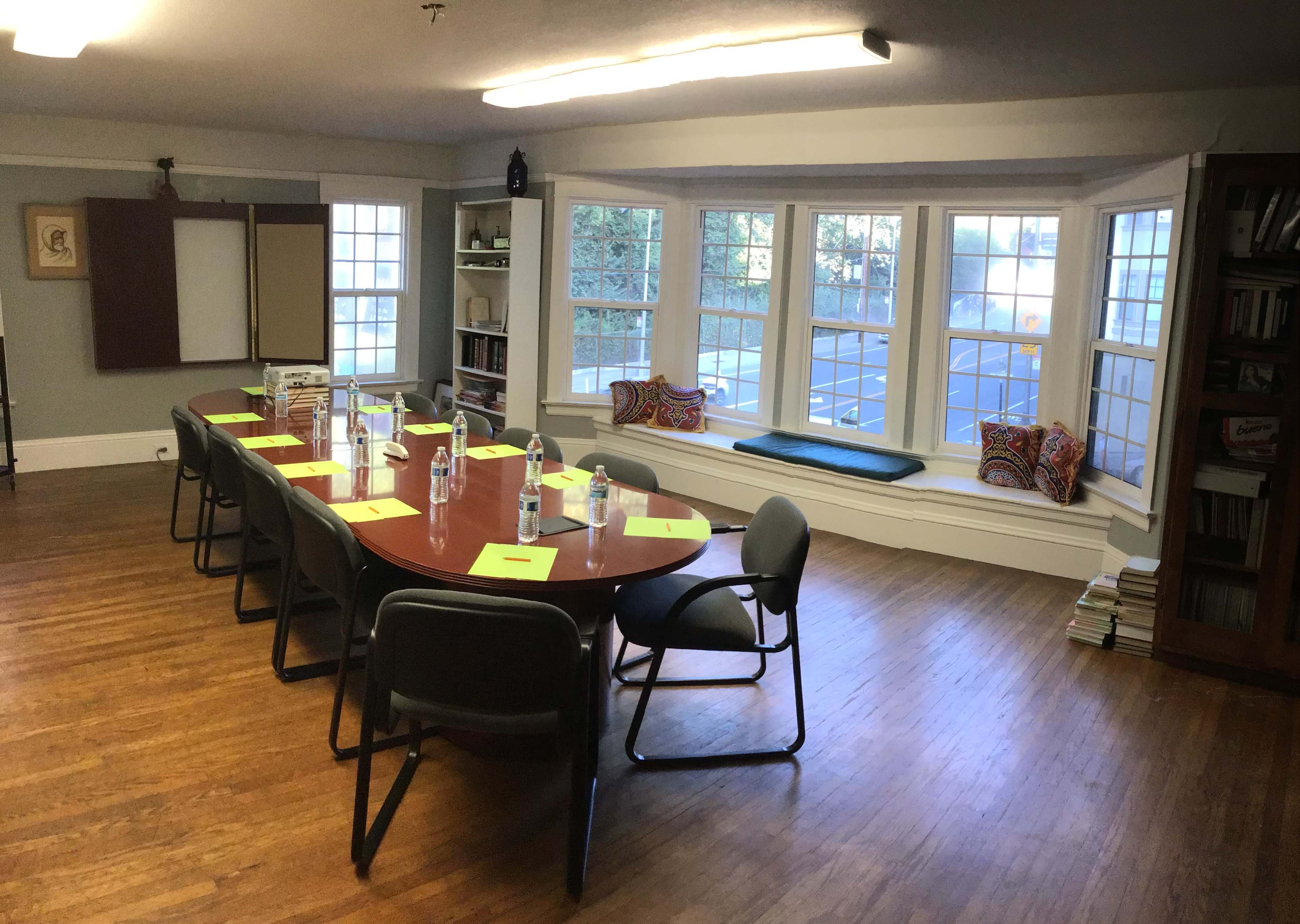 A rectangular conference table with chairs is arranged in a well-lit room featuring a window seat and shelves of books.