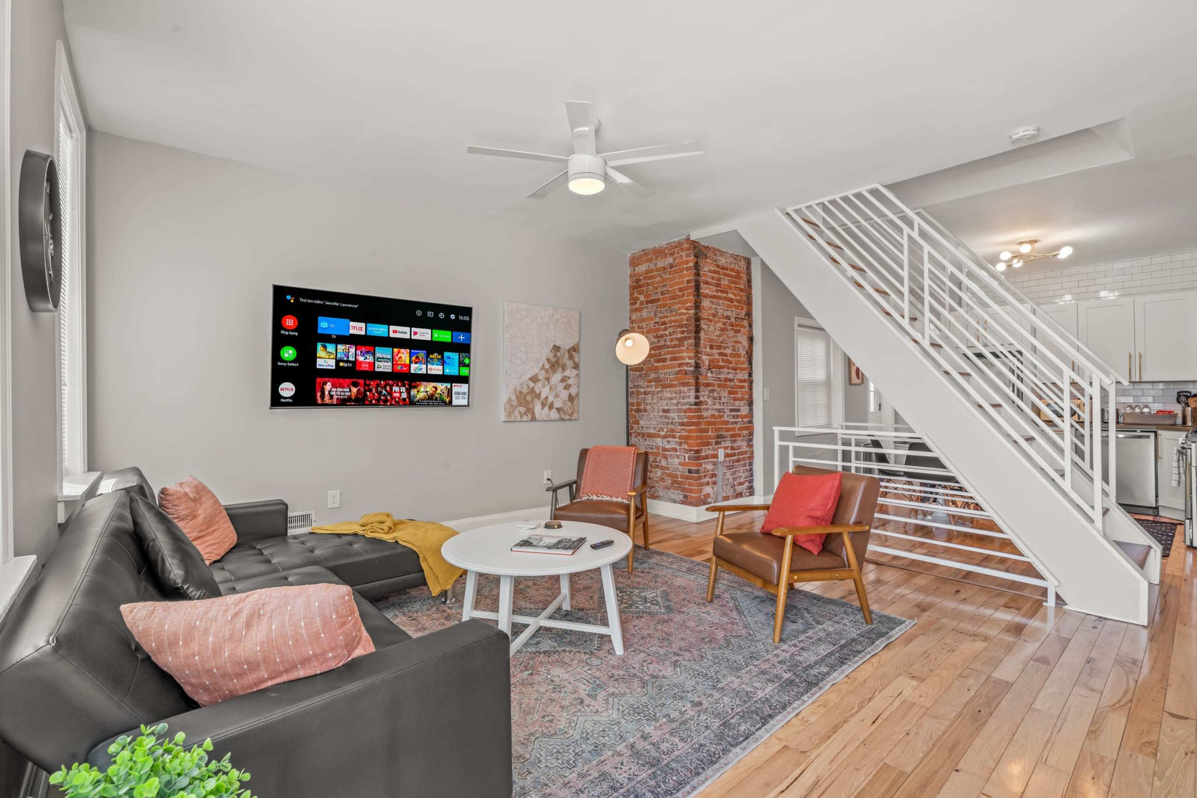 The living room features a modern design with a large wall-mounted TV, a sectional sofa, two accent chairs, a round coffee table, and a staircase leading to the upper level, complemented by exposed brick accents.
