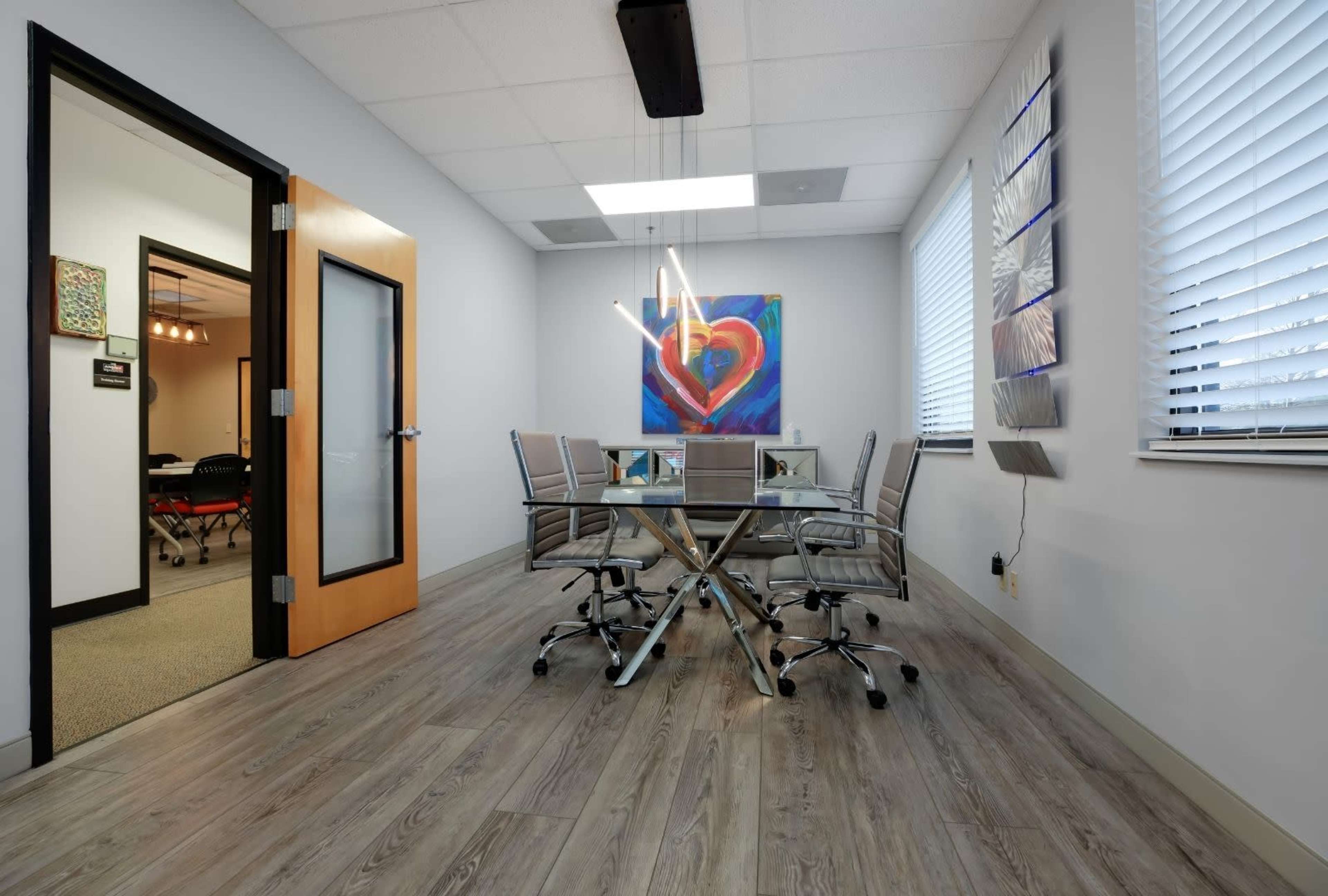 A conference room features a glass table surrounded by six chairs, with a colorful heart painting on the wall and a doorway leading to another office.