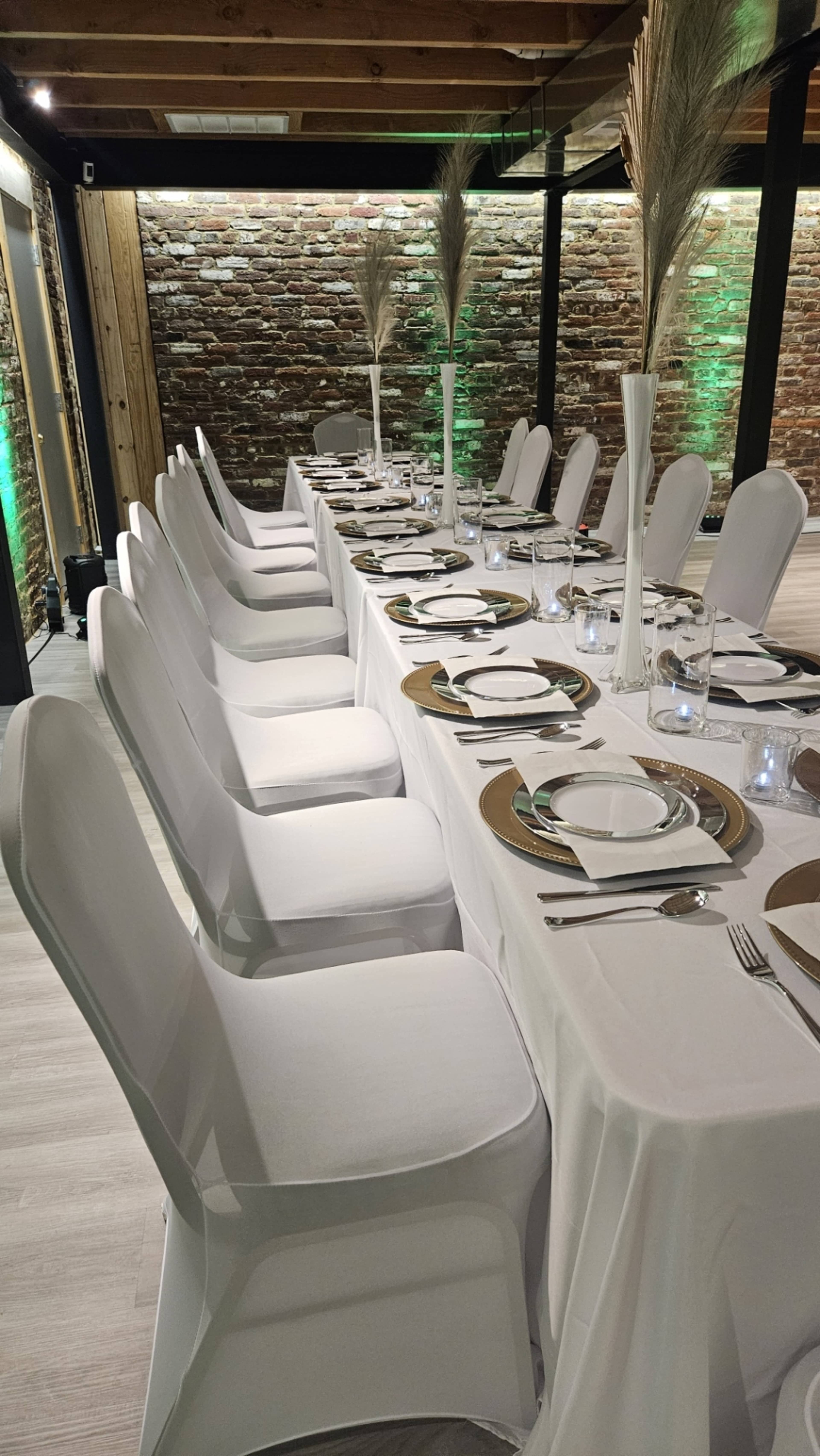 A long dining table set with white tablecloths and chairs, featuring plates, glasses, and decorative centerpieces, is arranged in a room with exposed brick walls and green lighting.