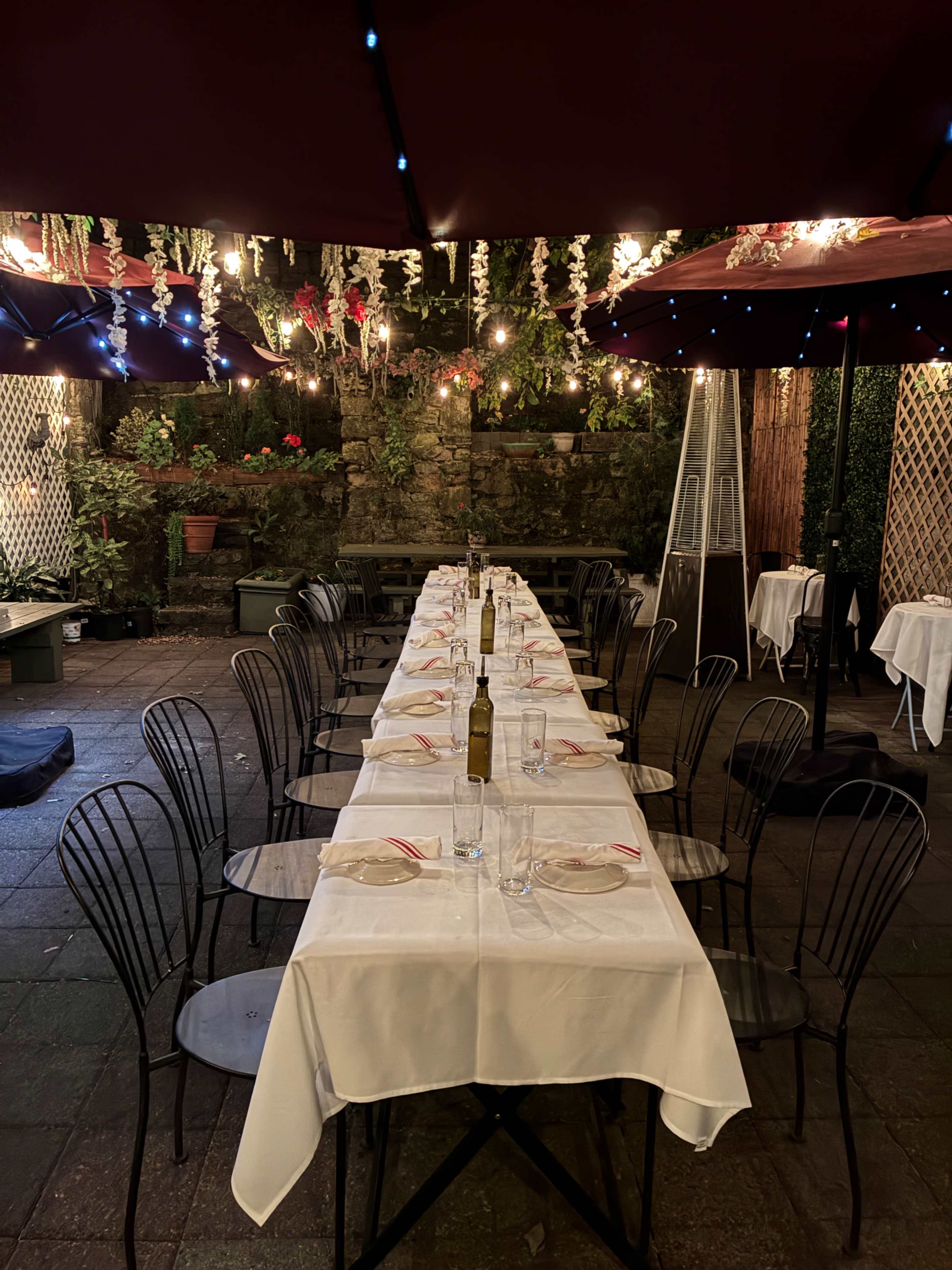 A long, elegantly set dining table with white tablecloths is arranged outdoors under red umbrellas, surrounded by greenery and string lights.