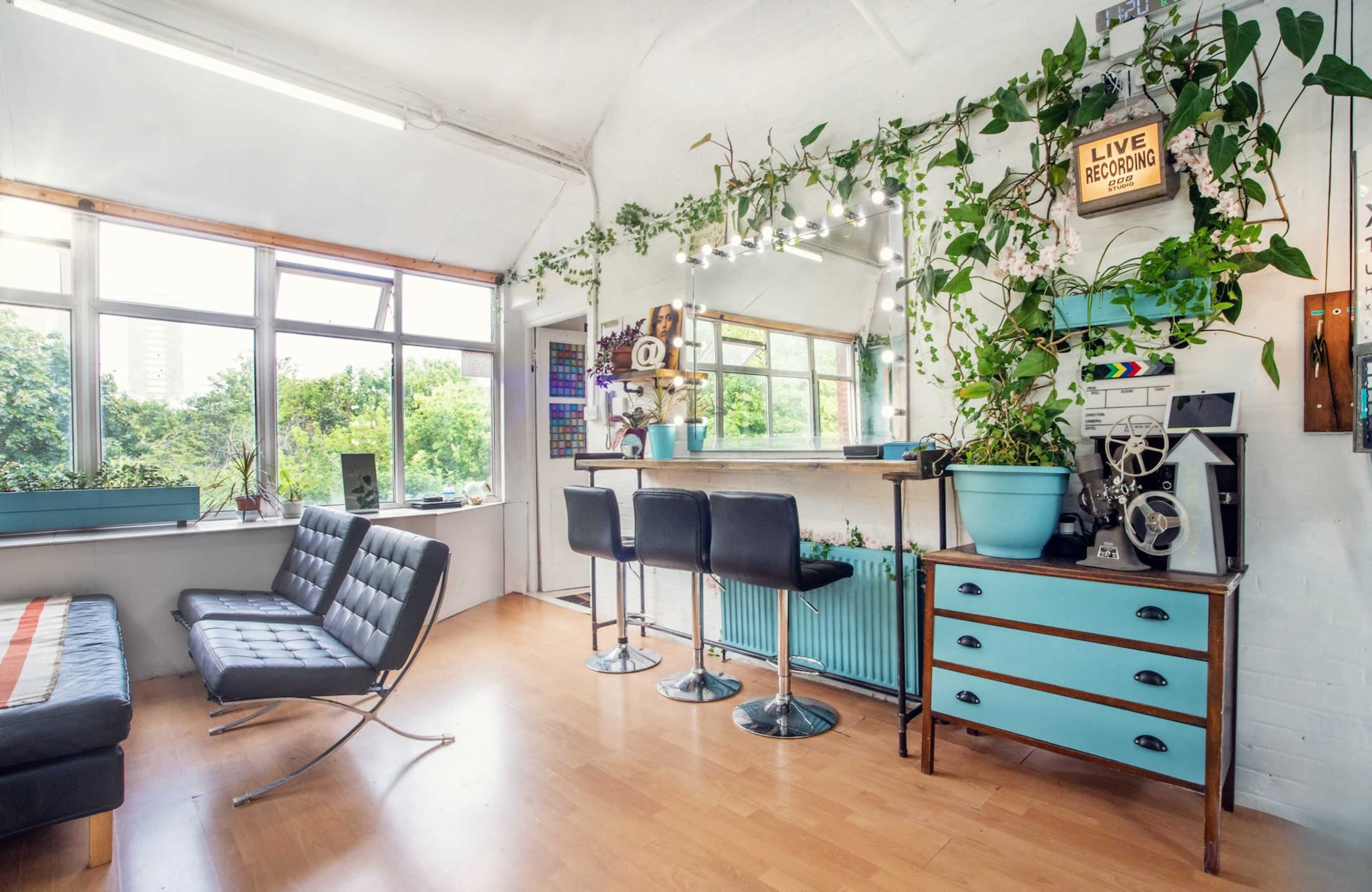 A bright, modern recording studio with large windows, a sleek sofa, bar stools, and greenery decor.