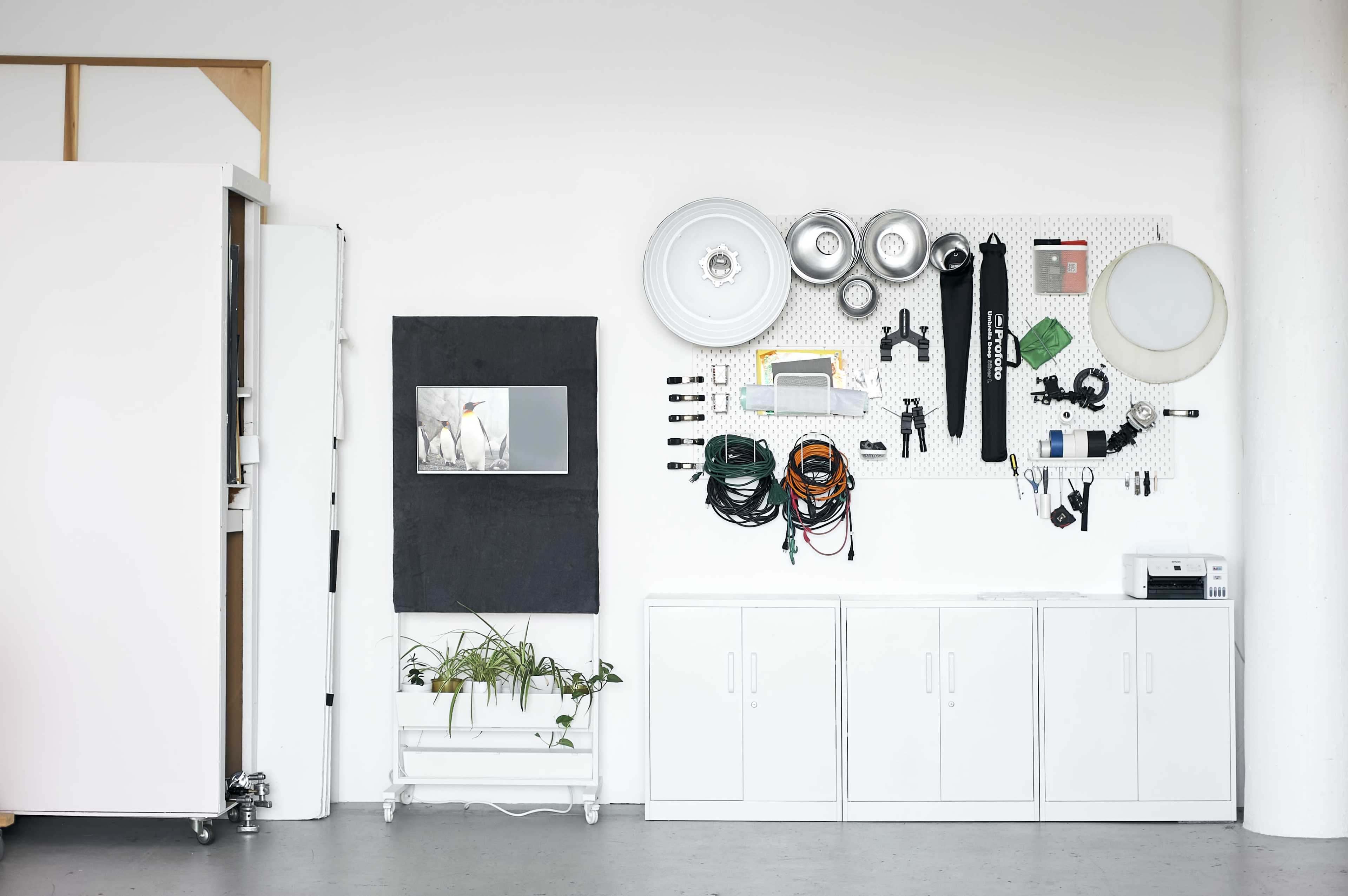 The image shows a neatly organized workshop wall featuring various tools, cables, and equipment mounted alongside a small television screen and a potted plant below.