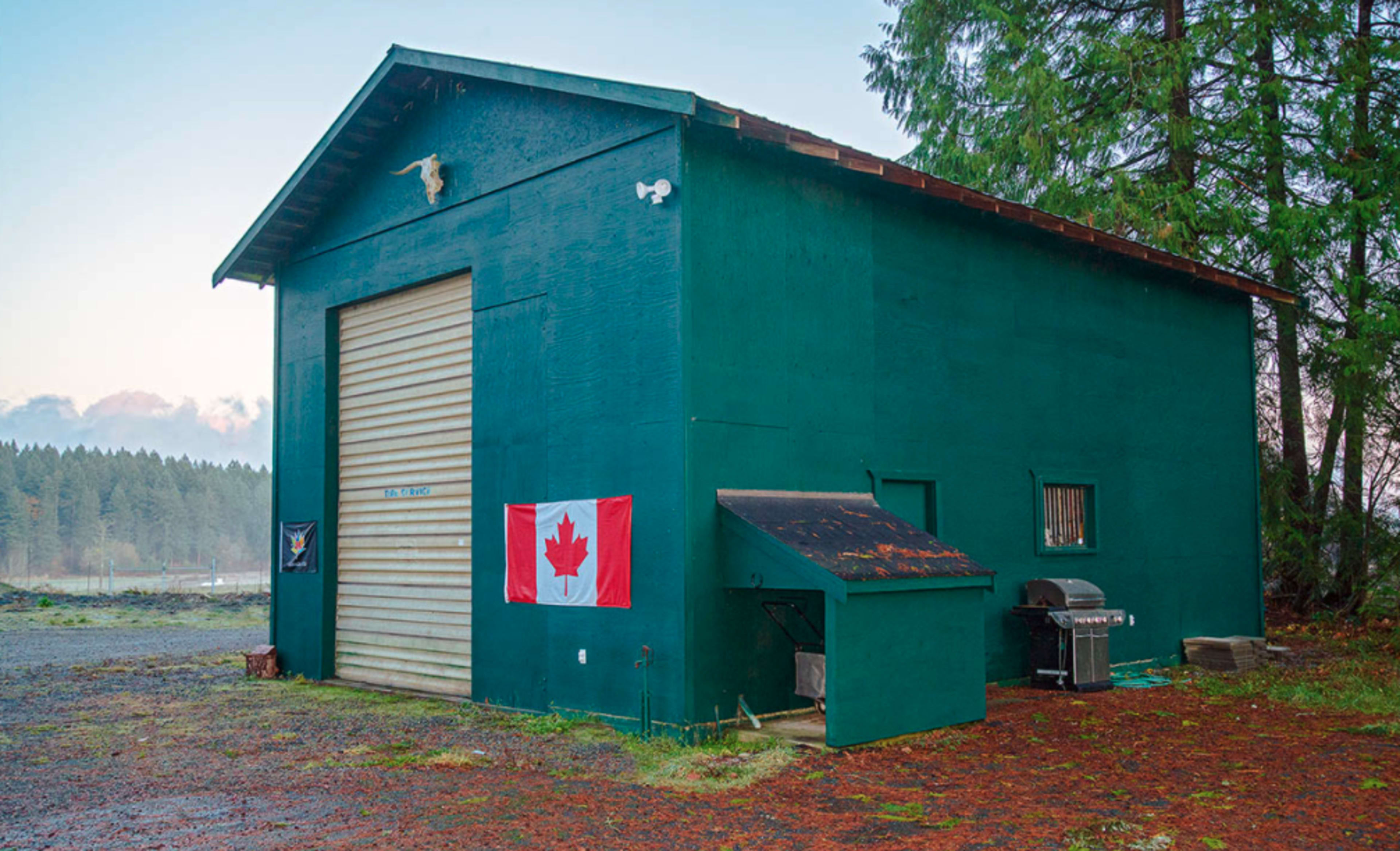 A large green building features a roll-up door, a Canadian flag hanging on the side, and a barbecue grill next to a small shed.