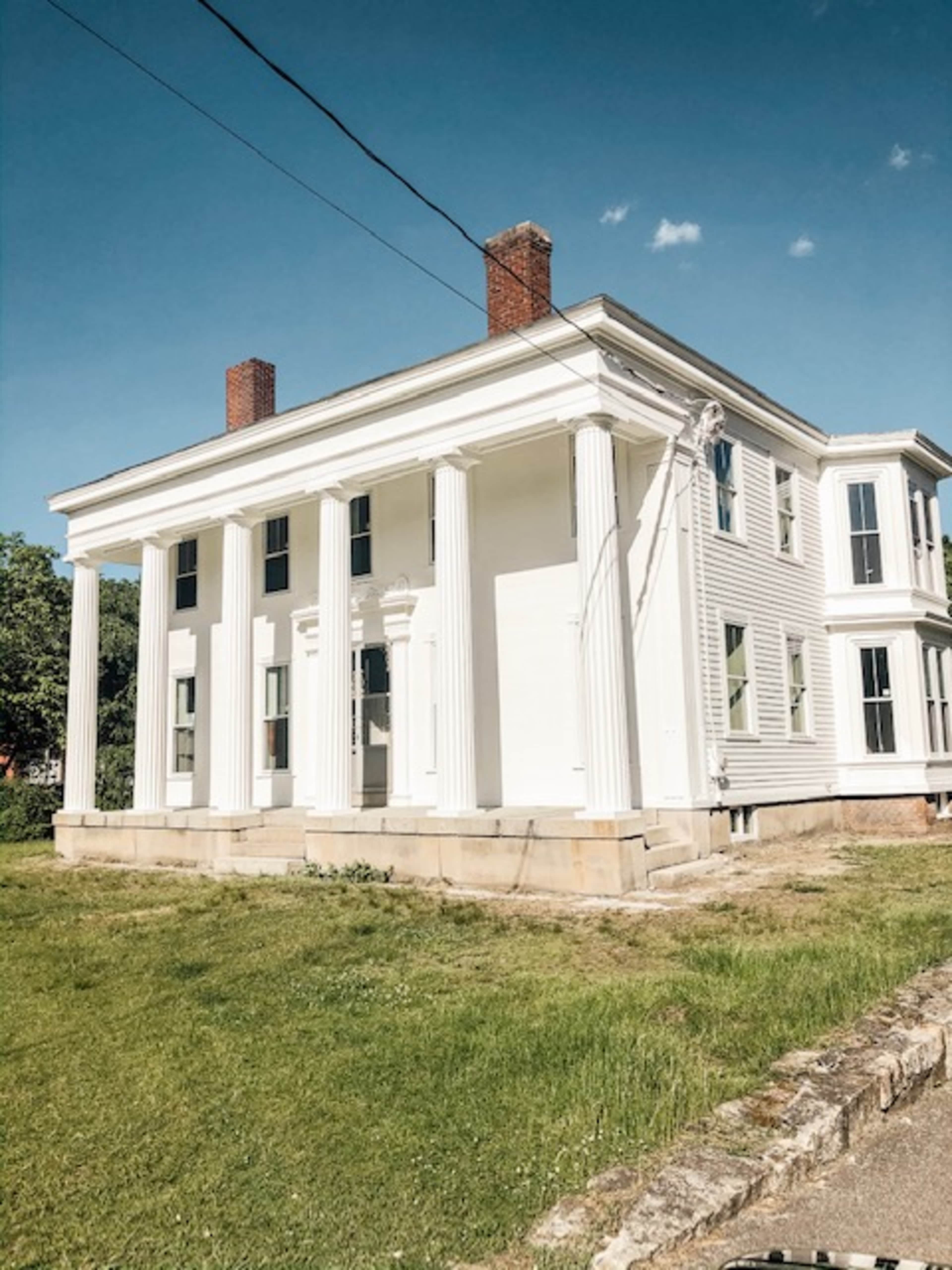 The image shows a large, white, two-story house with four columns at the front, set on a grassy lawn.