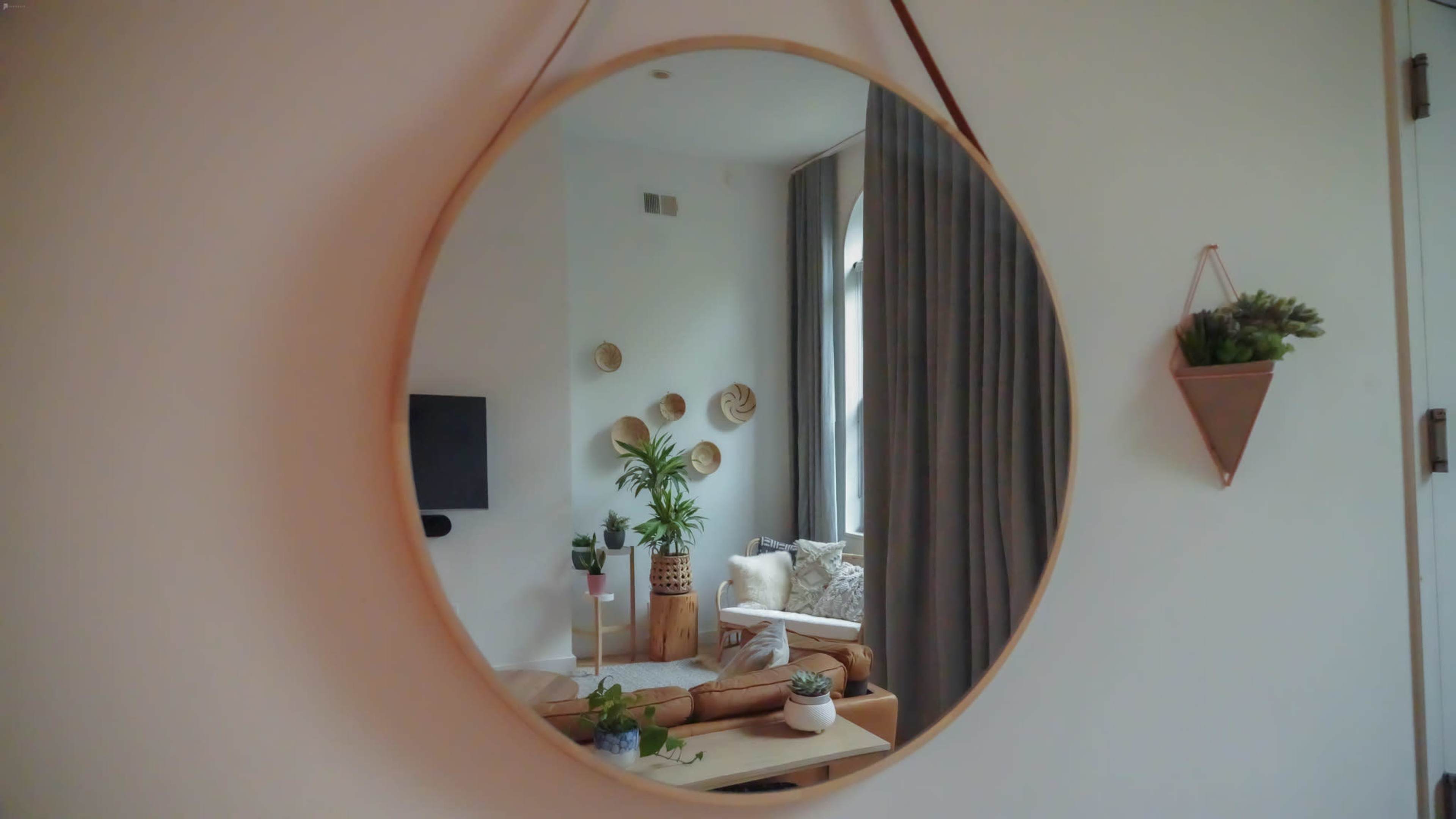 A round mirror hangs on a wall, reflecting a cozy living room with plants, a television, and decorative items.