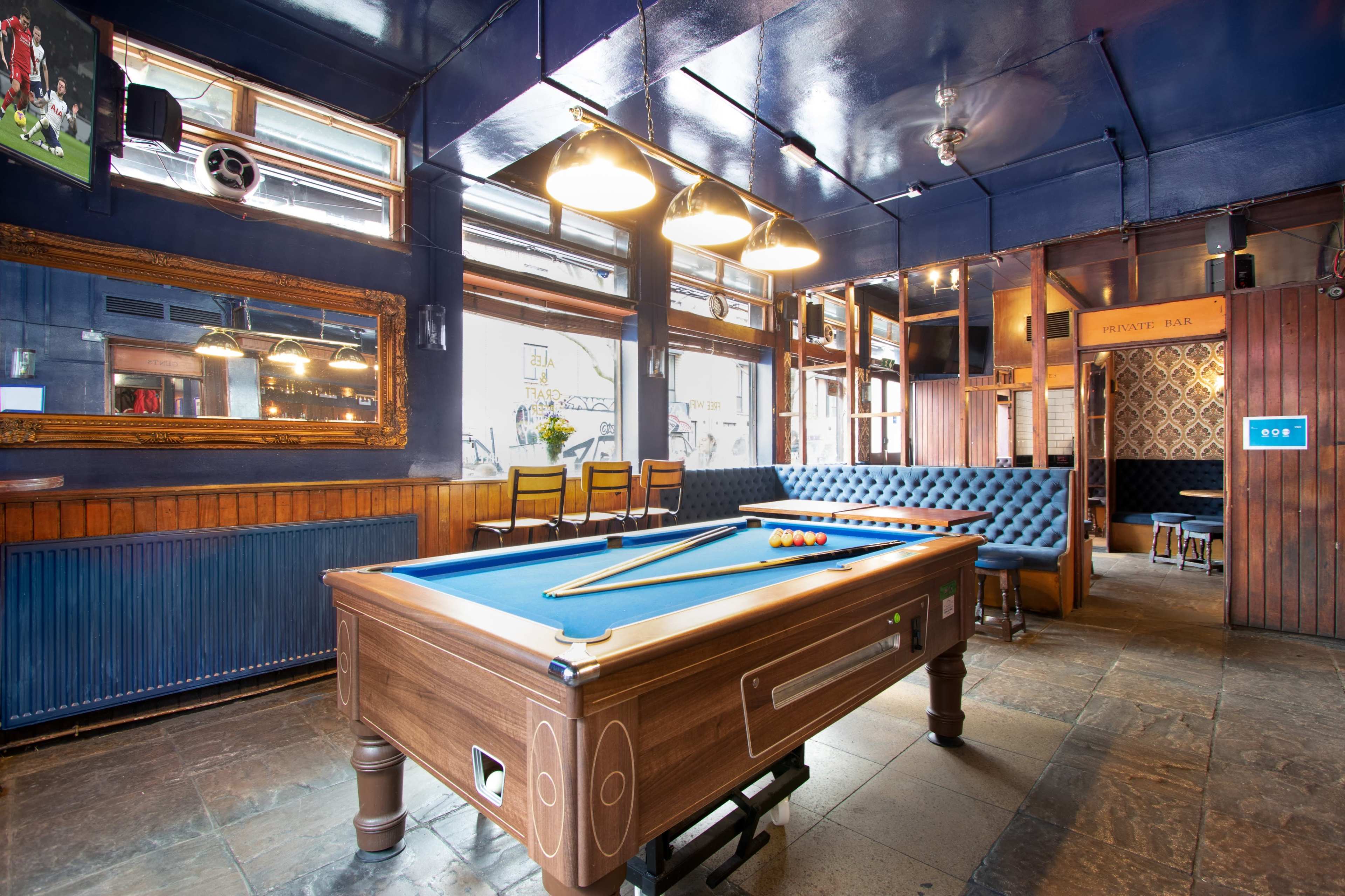 The image shows a bar interior with a billiards table, seating area, and large windows allowing natural light to enter.