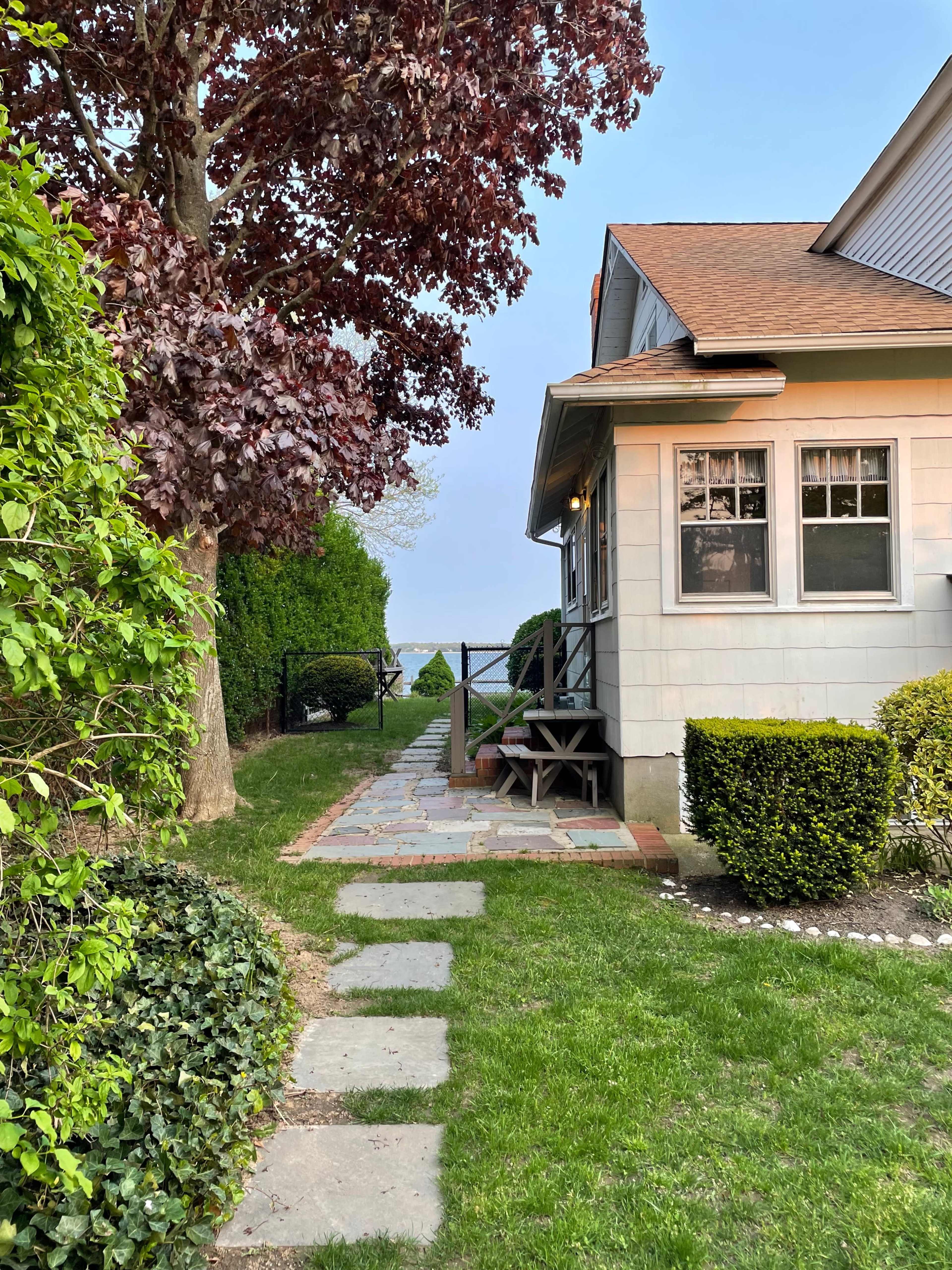 A stone pathway leads from a house to a wooden deck overlooking a body of water, bordered by green shrubs and trees.