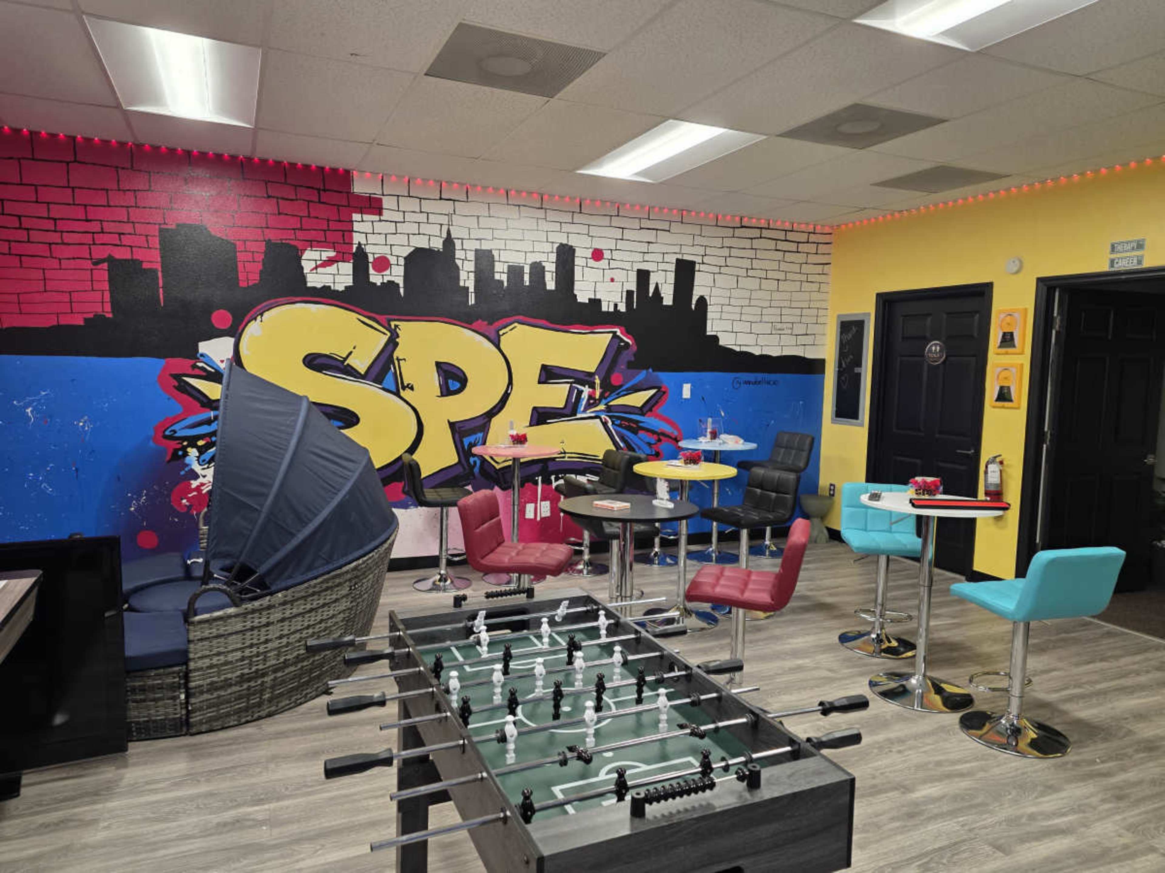The image shows a modern lounge area with colorful seating, a foosball table, and a vibrant painted mural on the wall.
