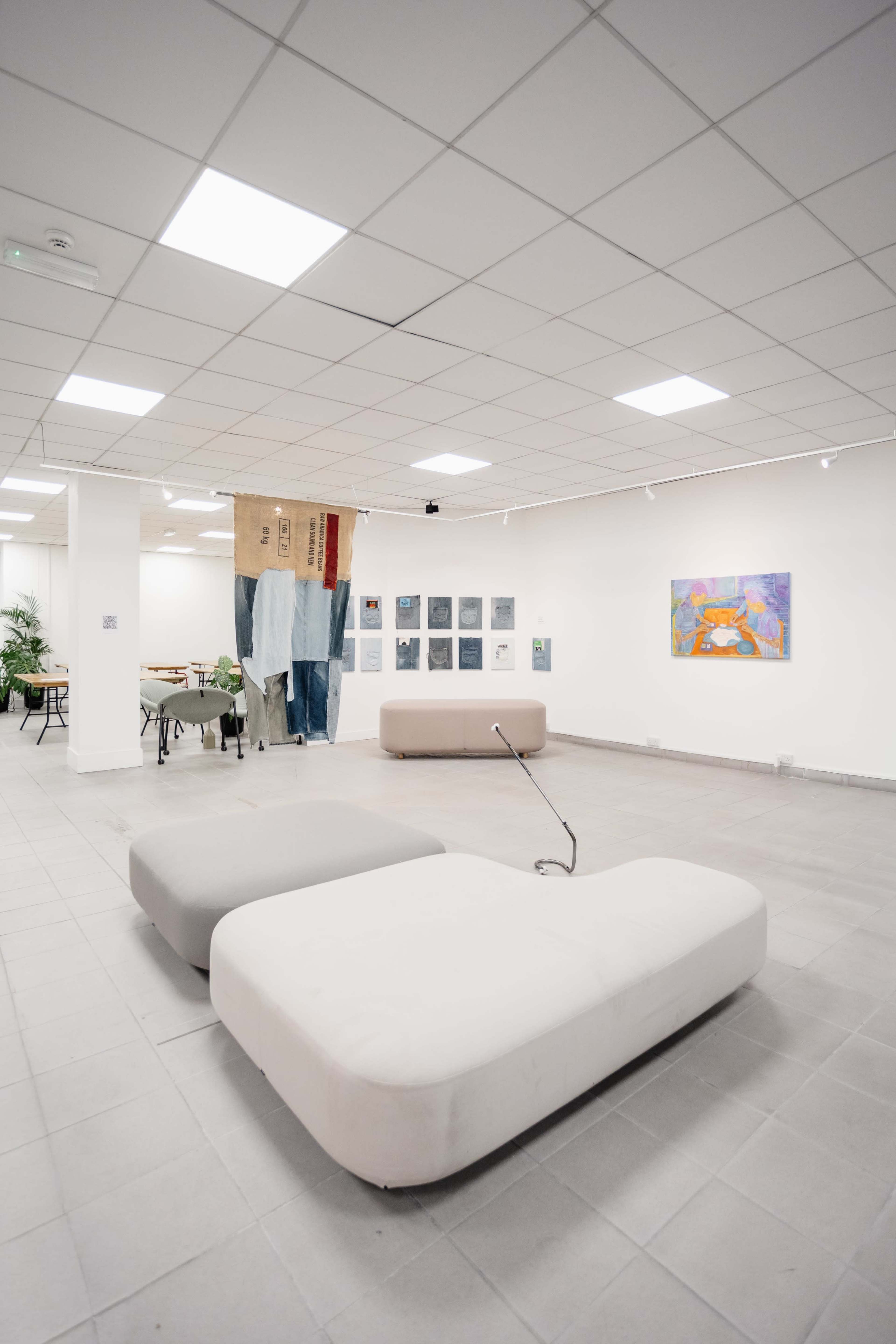 The image shows a spacious gallery with minimalist seating, an art installation featuring draped fabric, and various artworks displayed on the walls.