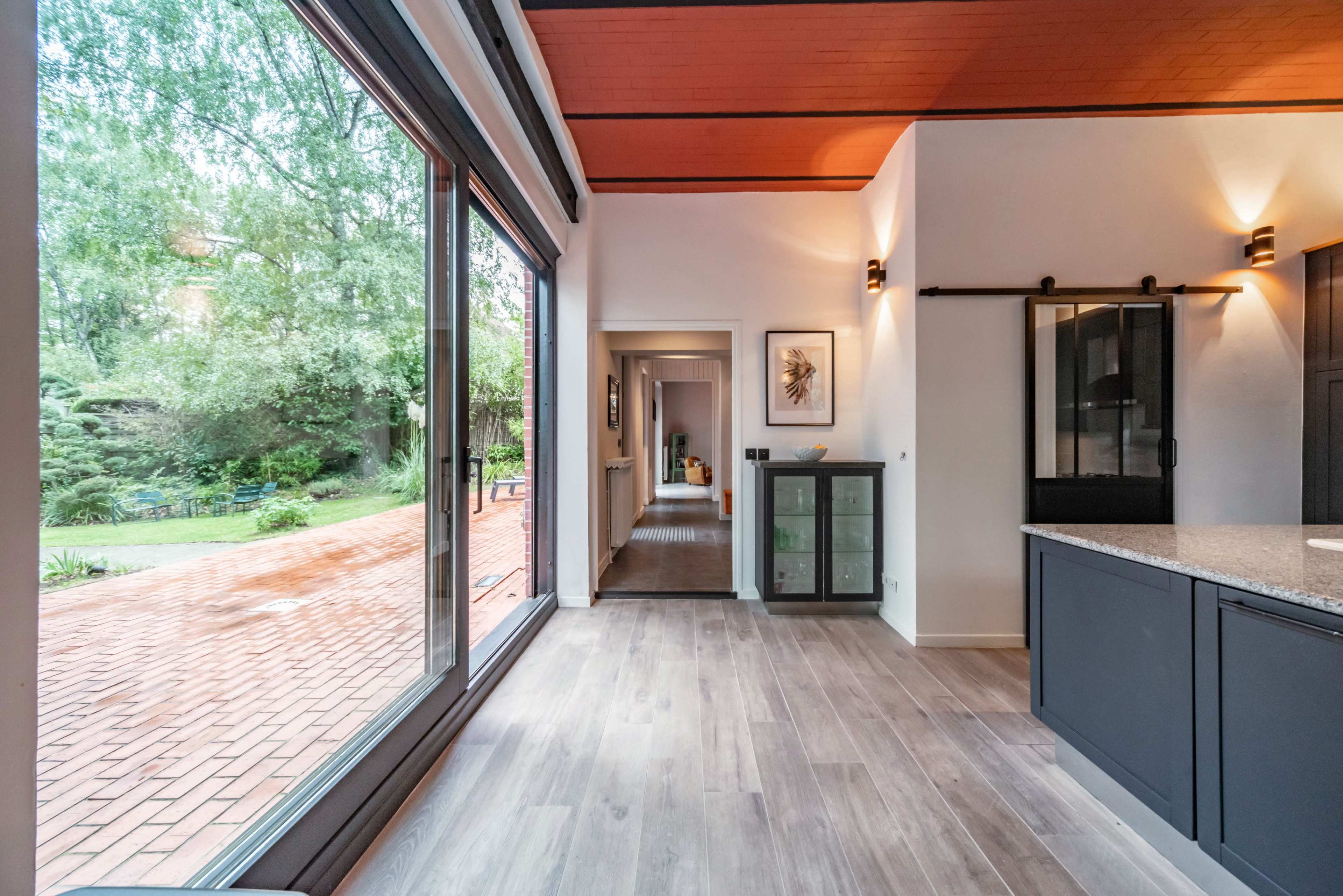 A modern kitchen area features large windows overlooking a garden, with a hallway leading to other rooms in the background.